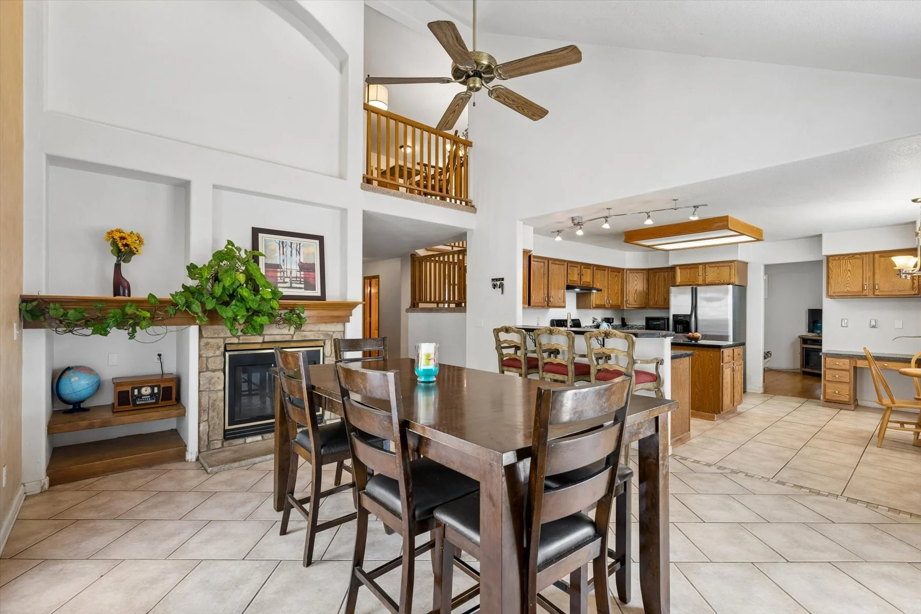 Open-concept kitchen and dining area with hardwood and tiled flooring, featuring a dining table with six wooden chairs, a kitchen with wooden cabinets, stainless steel appliances, a breakfast bar with four chairs, and a fireplace with a stone surround and mantel decorated with a vase of flowers and a framed picture, with a staircase and a ceiling fan visible in the background.