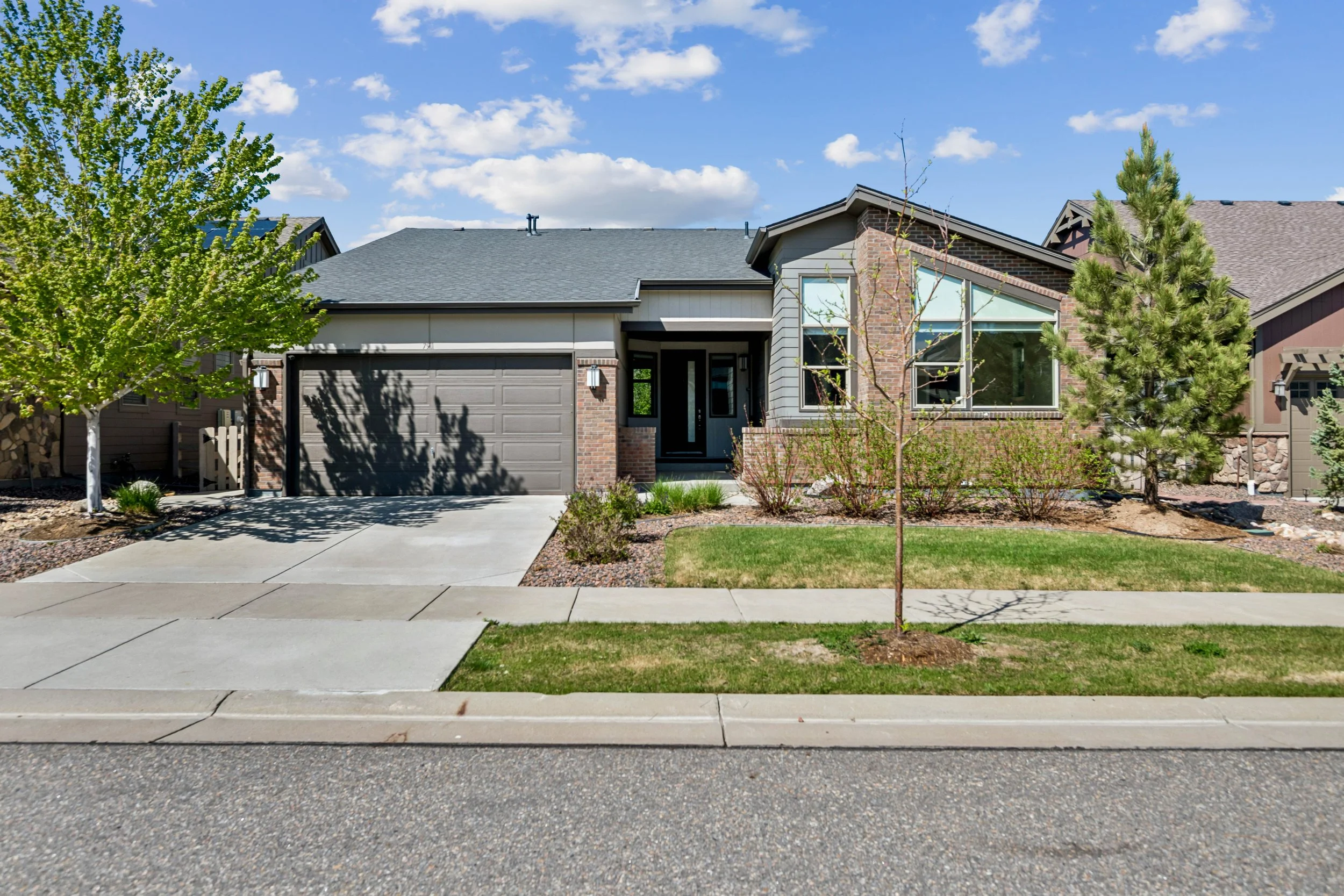 Front view of a modern suburban house with a two-car garage, driveway, landscaped yard with trees and shrubs, under a blue sky with some clouds.
