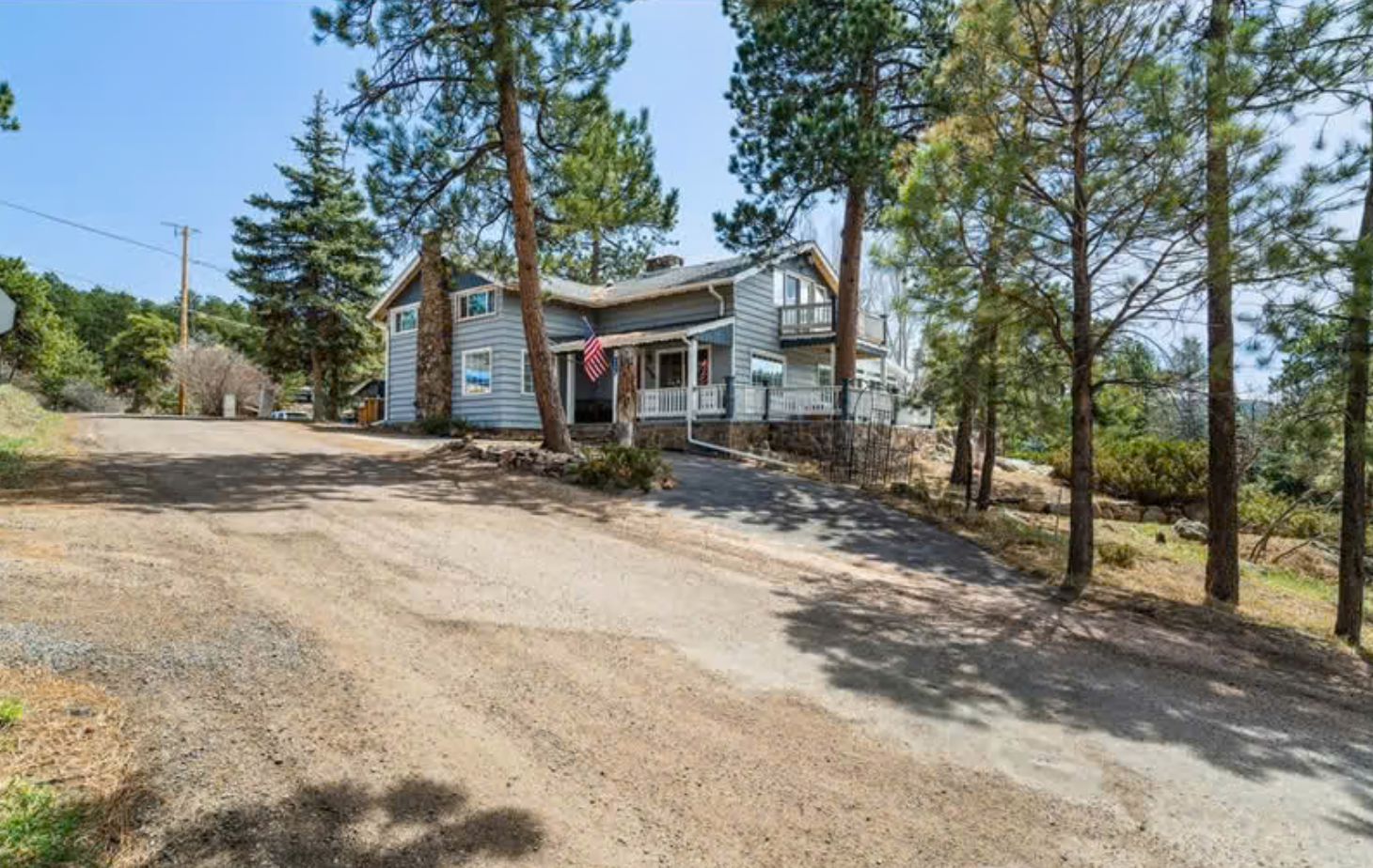 A house with blue siding and a white porch surrounded by tall pine trees, with a gravel driveway leading up to it on a sunny day.