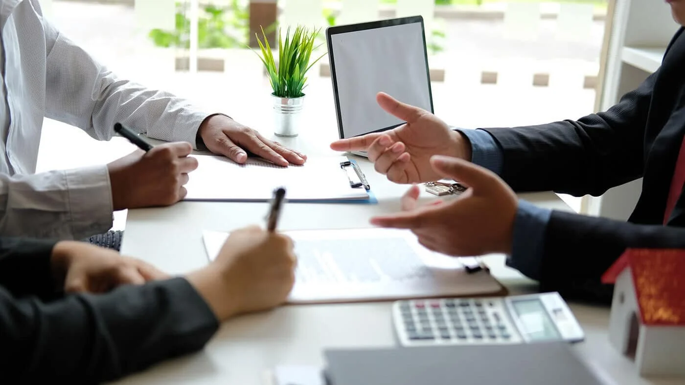 People in business suits having a discussion at a table, with a laptop, documents, a calculator, and a small house model on the table.