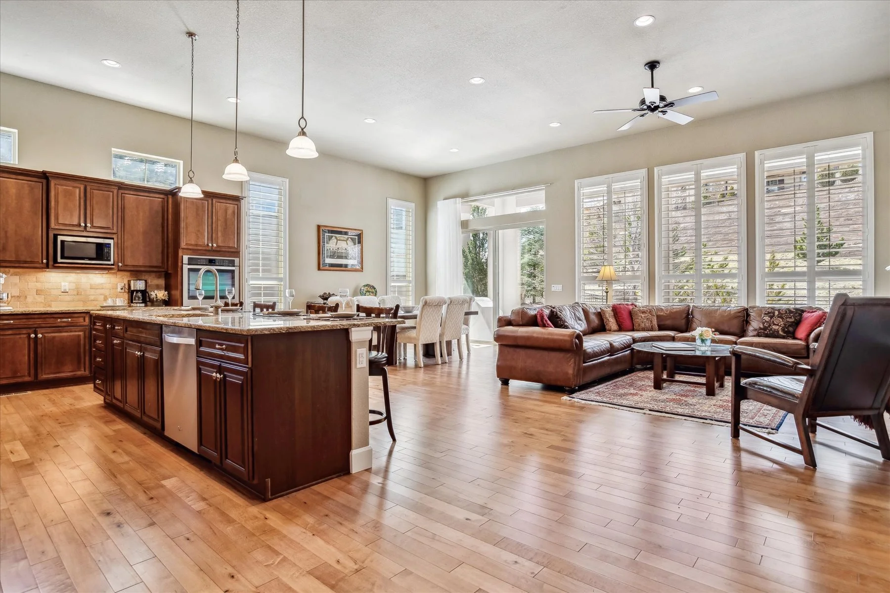 Open-concept living room and kitchen with hardwood floors, large windows with white blinds, a brown leather sectional sofa with pillows, a glass coffee table, a dining table with chairs, and wooden kitchen cabinets, ceiling fan, and pendant lights.