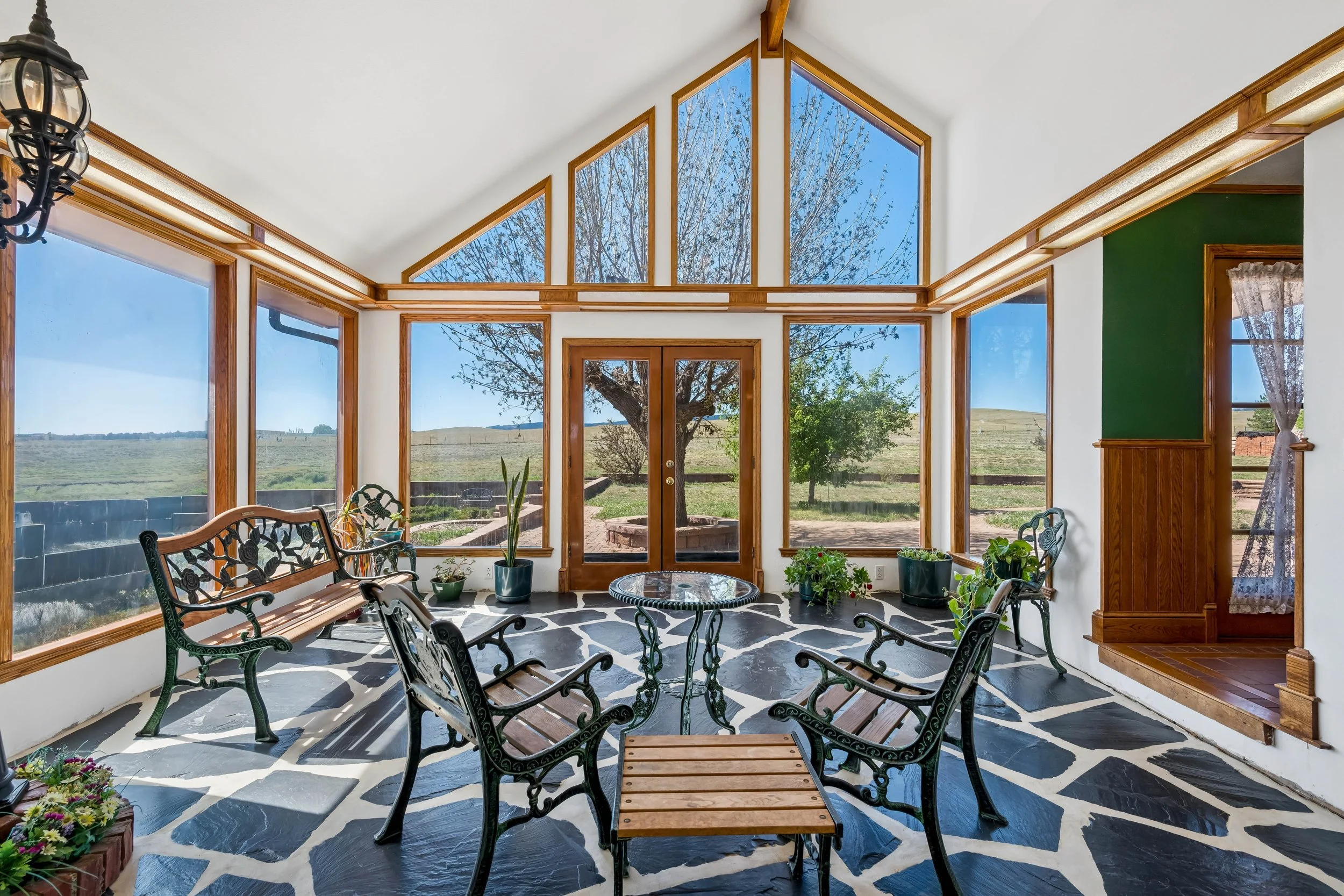 Interior view of a sunroom with floor-to-ceiling windows, black and white patterned floor, wrought iron furniture, and potted plants, overlooking a rural landscape with trees and open fields.