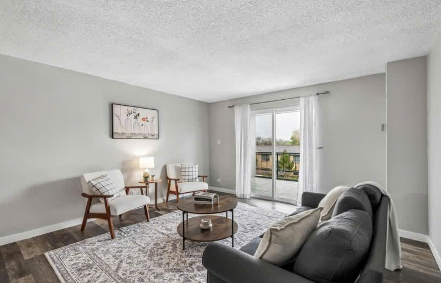 Living room with gray walls, a sliding glass door leading to a balcony, a dark gray sofa with throw pillows, two white armchairs with wood arms, a round coffee table, a patterned rug, a wall art piece, and sheer white curtains.