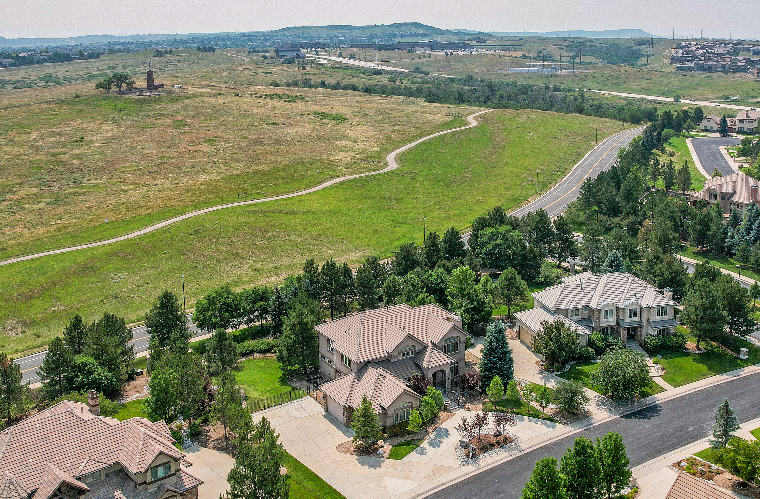 Aerial view of suburban neighborhood with houses, trees, a winding path, and open grassy hills in the background.