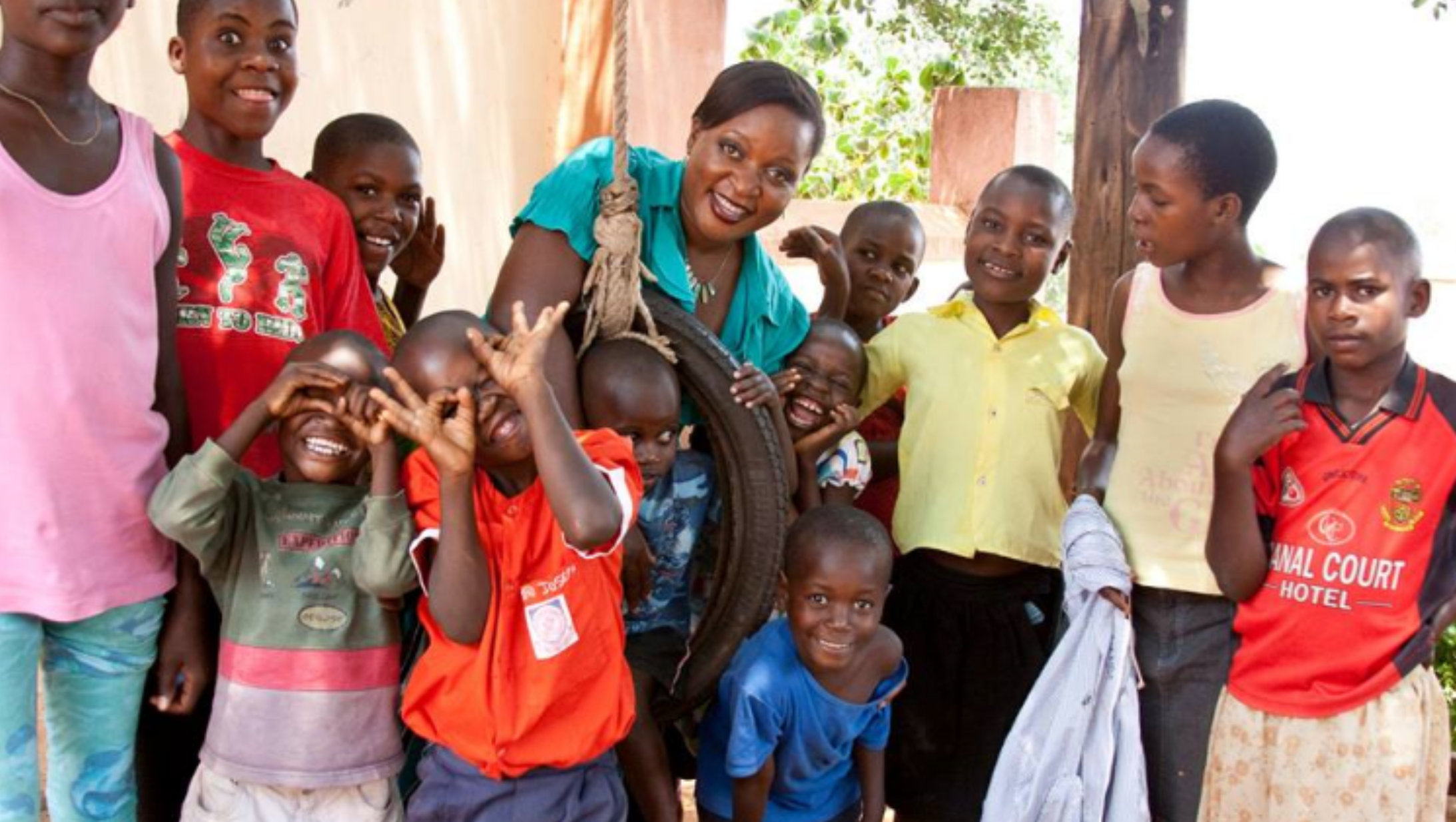 Group of children and a woman, some children are smiling and making playful gestures, with a snake wrapped around the woman. They are outside in a setting with trees and a wooden post visible in the background.