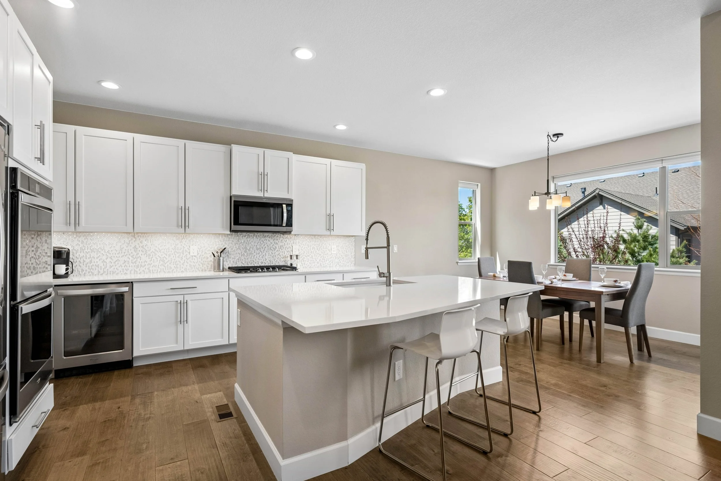 Modern kitchen with white cabinets, a kitchen island with bar stools, stainless steel appliances including a microwave and oven, a gas stove, a mosaic tile backsplash, and a dining area with a wooden table and chairs near large windows bringing in natural light.