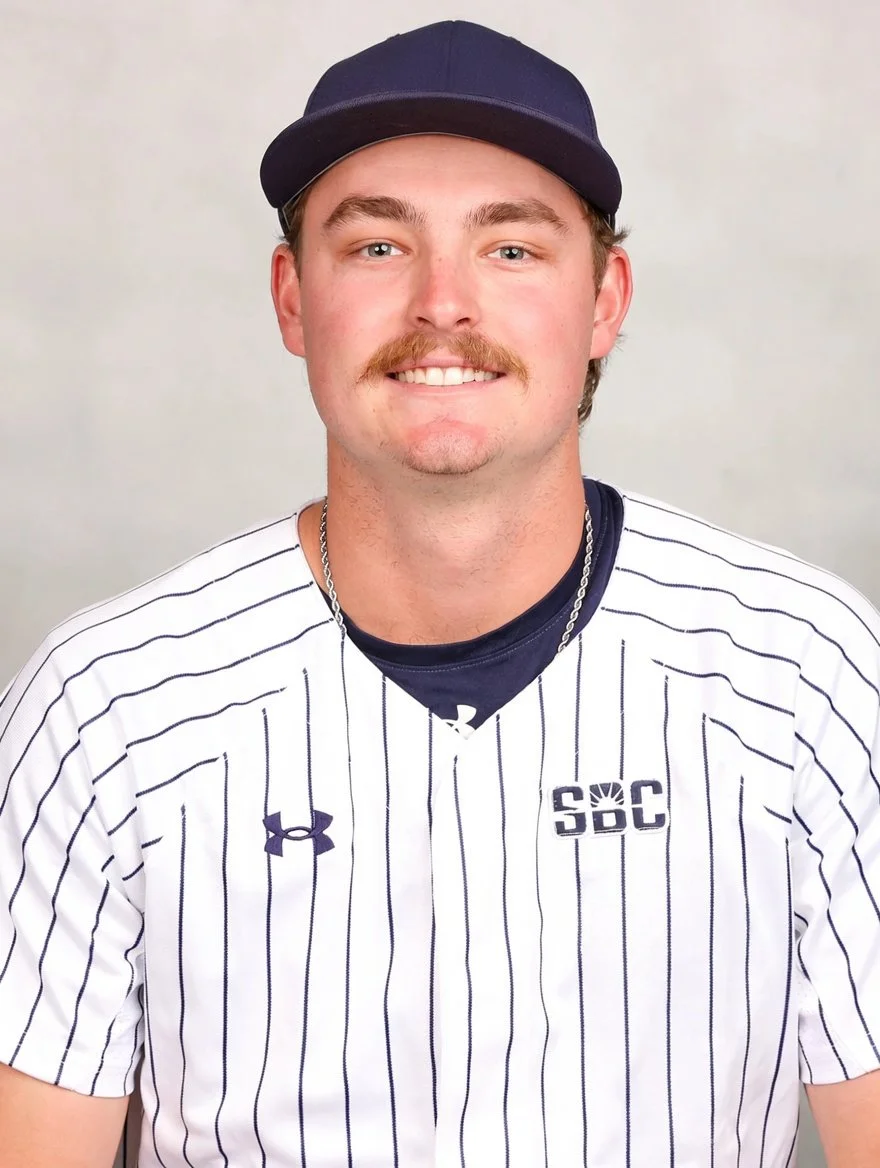 A young man with blond hair, blue eyes, and a mustache, wearing a navy cap, white baseball jersey with black pinstripes, and a chain necklace, smiling at the camera.