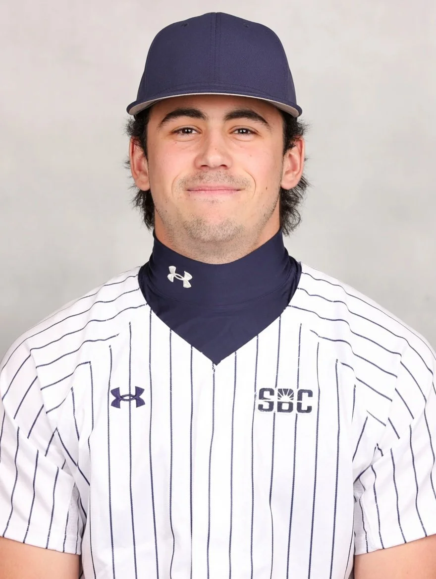 Portrait of a young man wearing a baseball uniform, navy cap, black neck gaiter, and smiling.