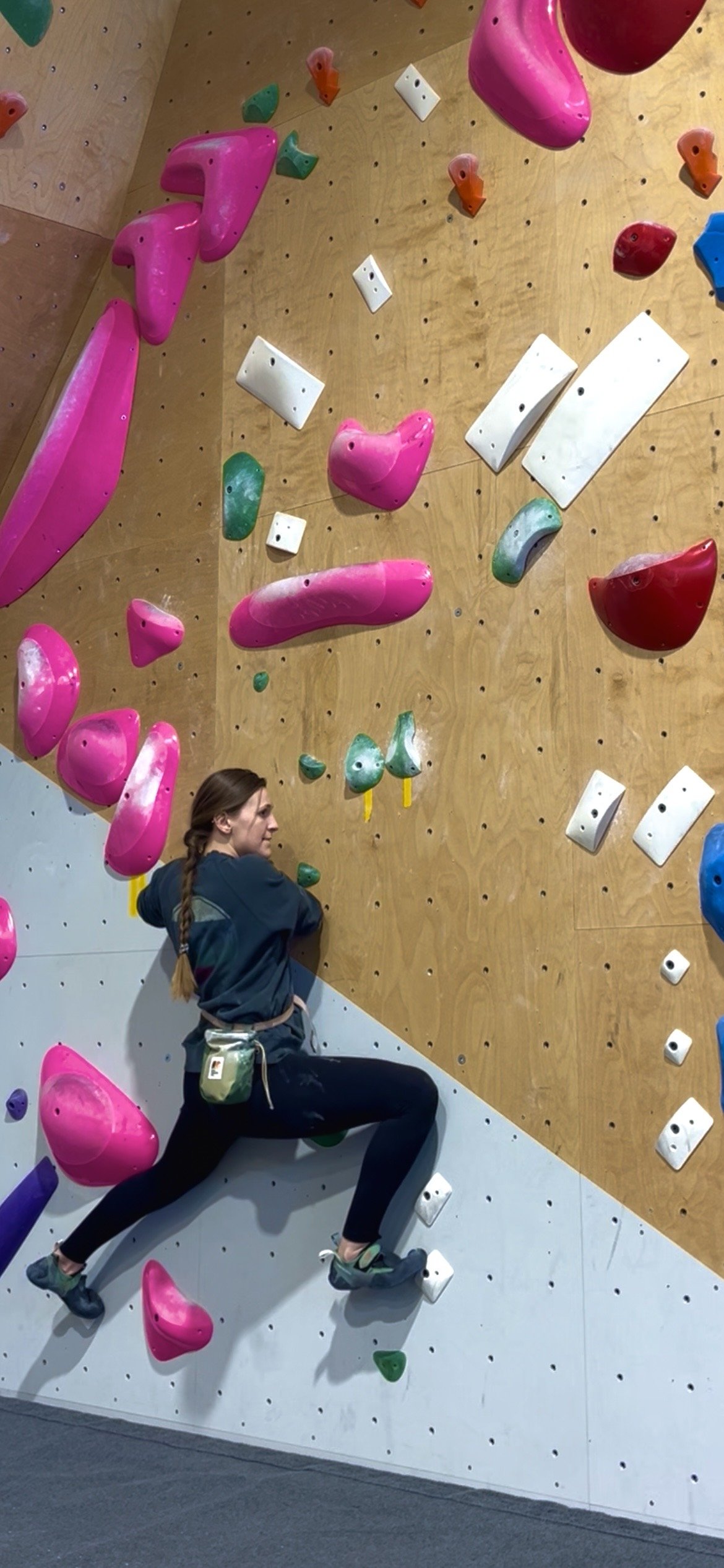 A woman climbing an indoor bouldering wall with colorful holds, including pink, green, white, red, purple, and blue, in a gym setting.