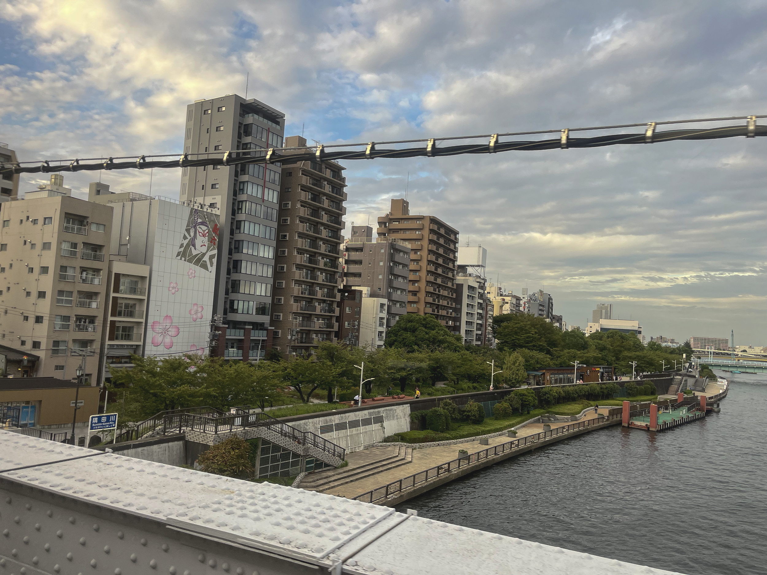 Cityscape with tall buildings along a river, partly cloudy sky, and trees at the riverbank, with a bridge rail in the foreground.