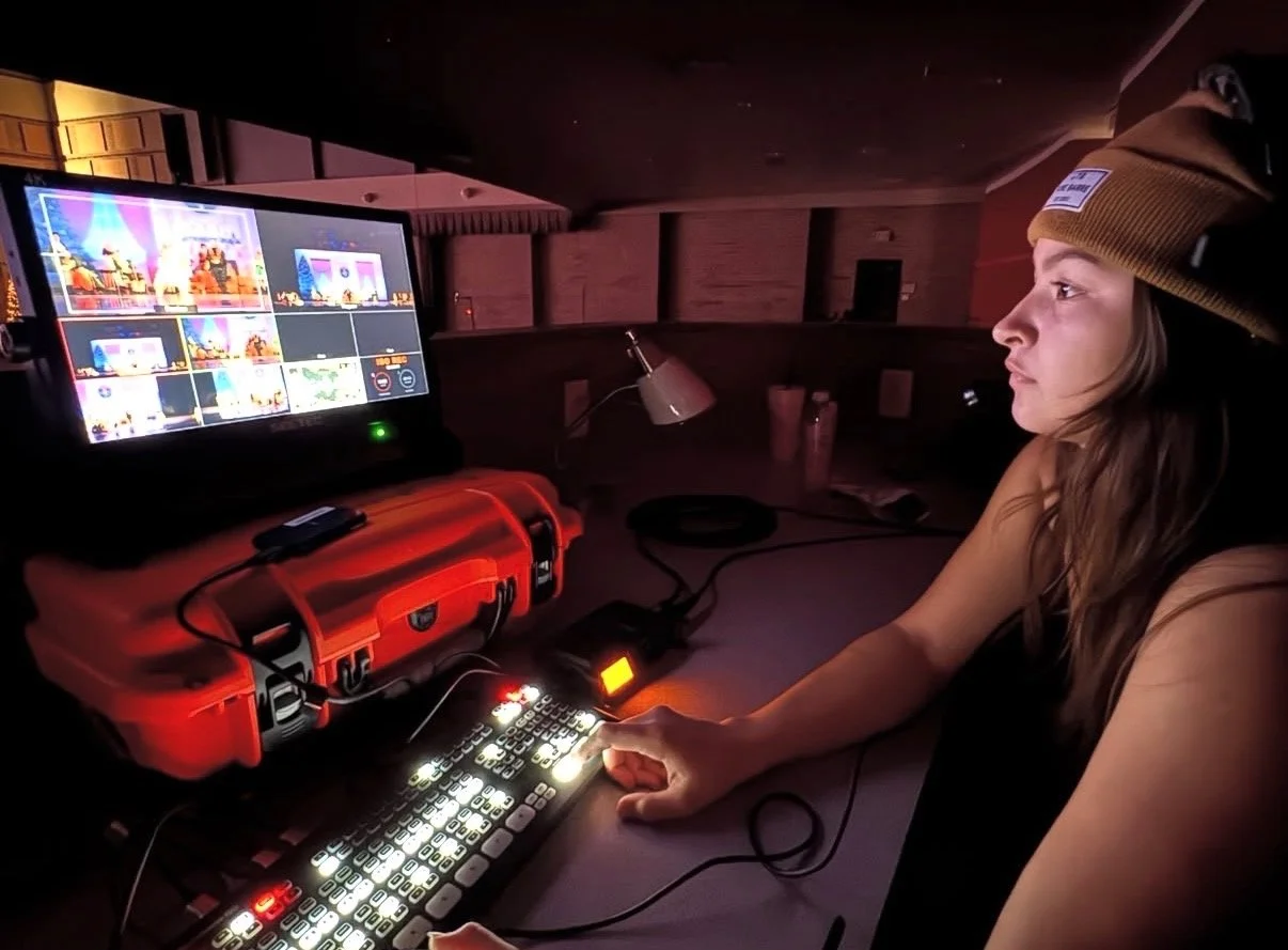 A woman in a brown beanie working at a video production control desk with a monitor displaying multiple video feeds.