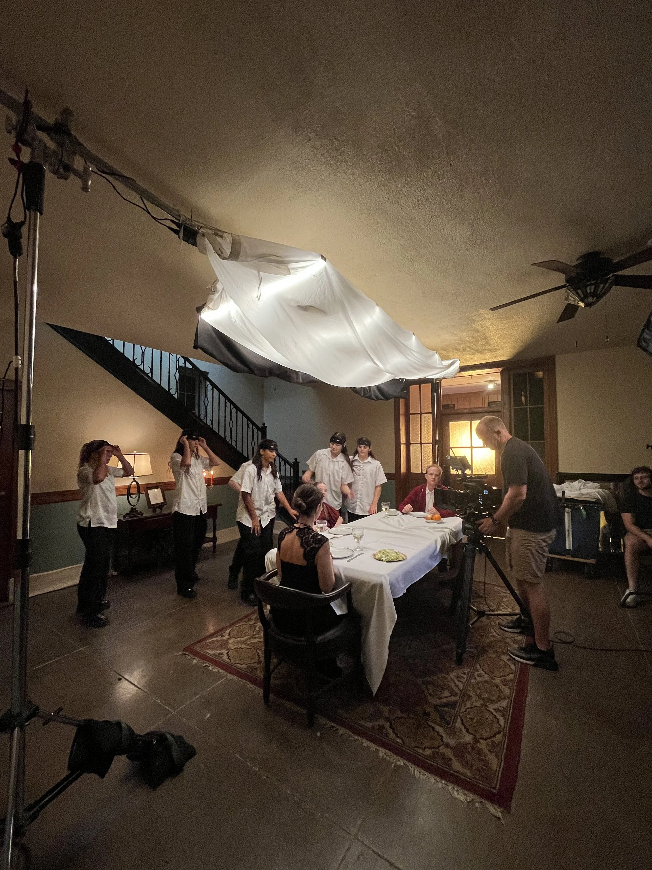 Behind the scenes of a film or photo shoot in a dining room, with crew members setting up lights and cameras, and people seated at a table.