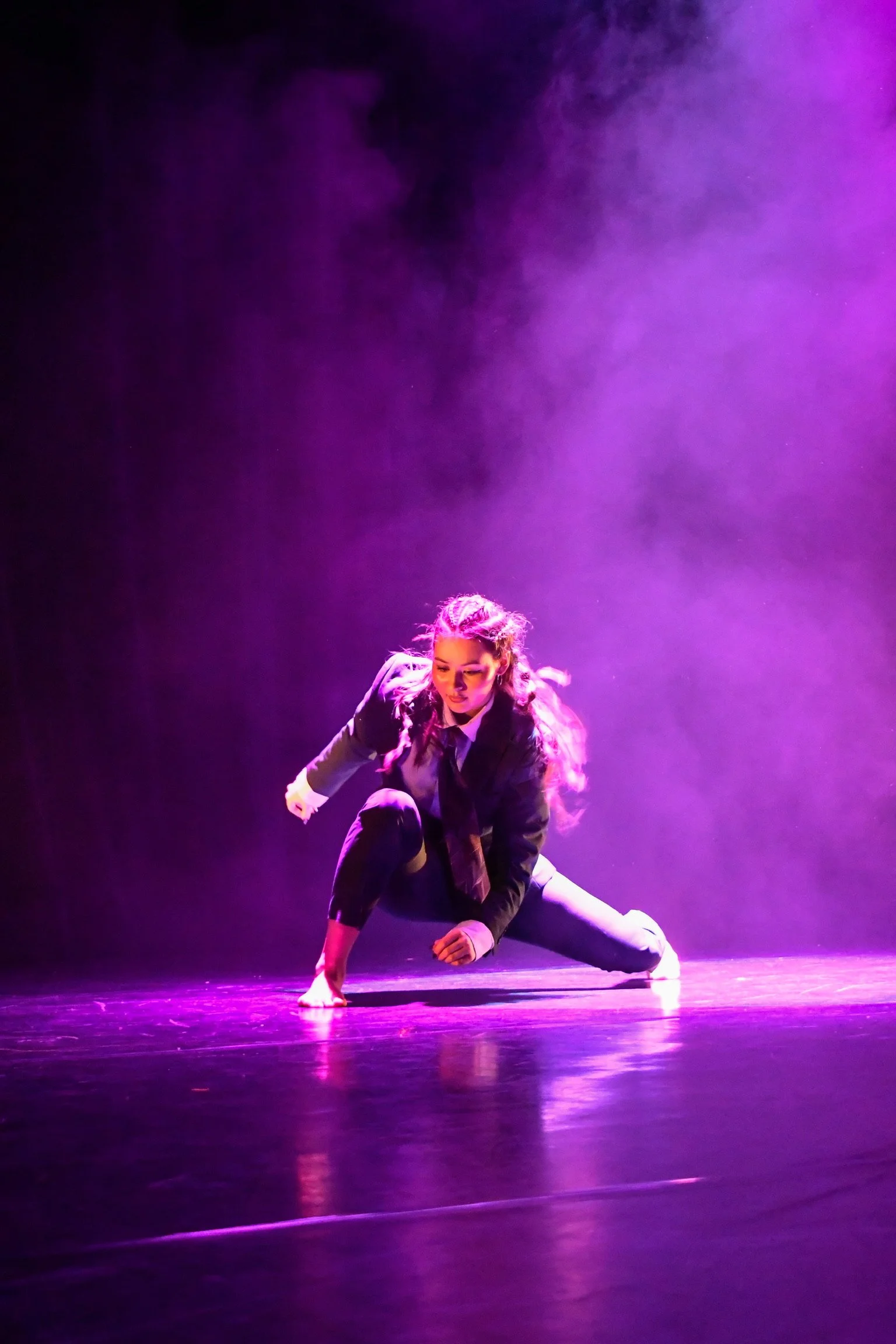 A female dancer dressed in a black suit performs a contemporary dance move on stage, illuminated by purple and pink stage lights, with smoky atmosphere in the background.