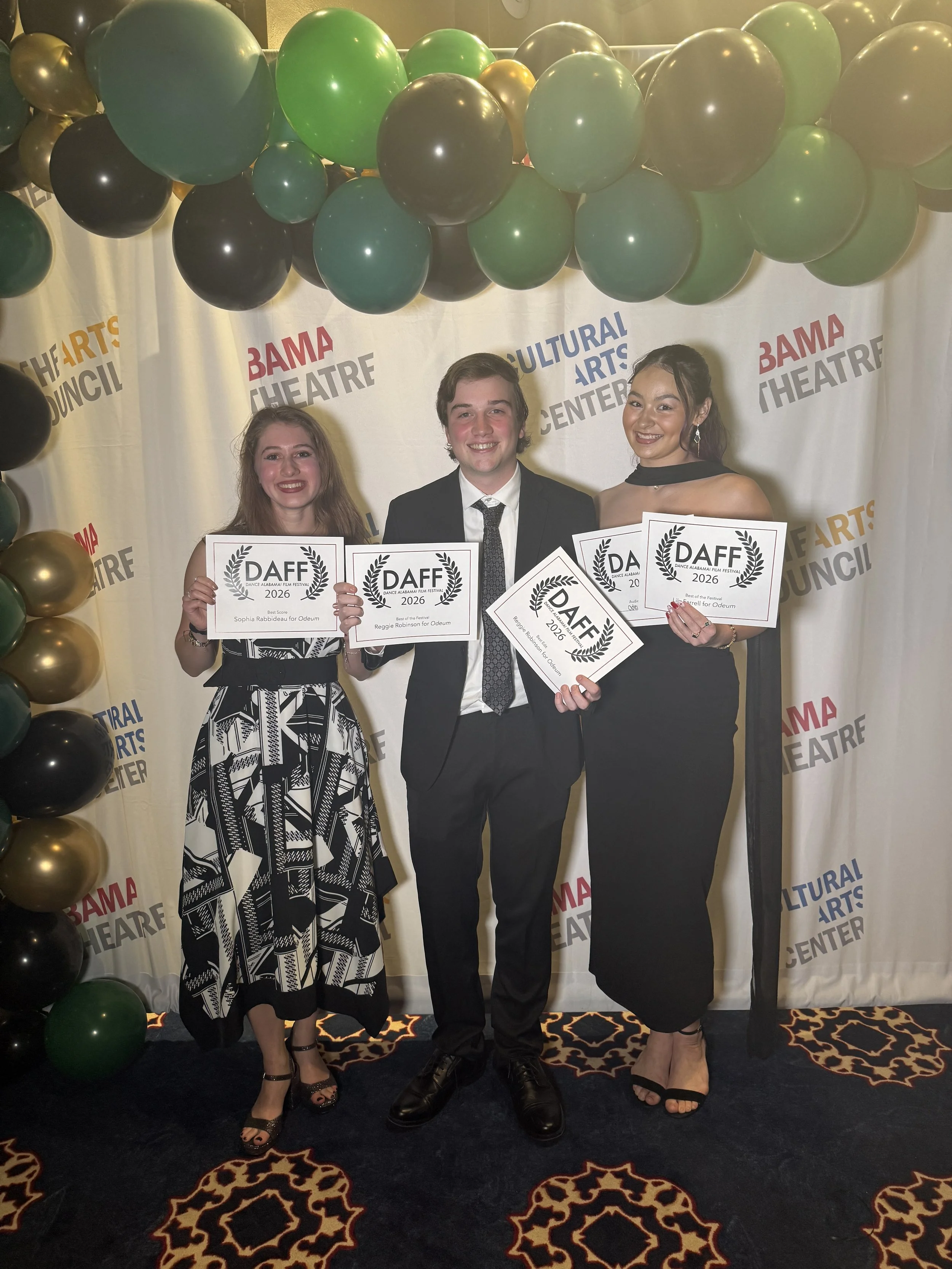 Three young adults smiling and holding awards at a cultural arts center with black and gold balloons and a decorative carpet.
