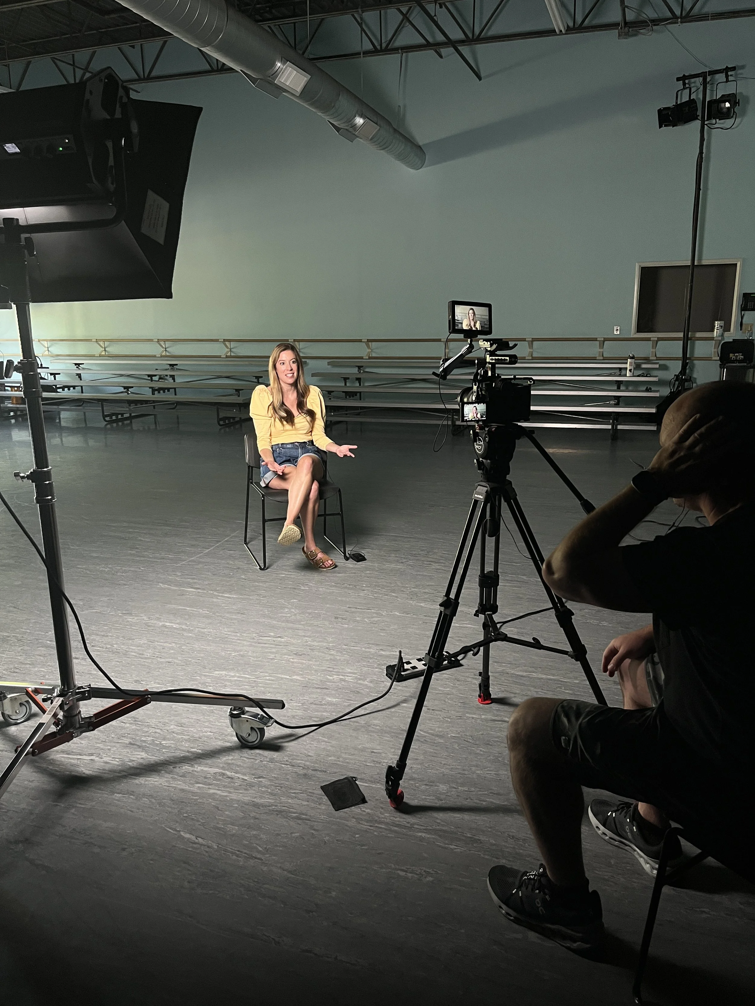 A woman with long hair wearing a yellow top and denim skirt is sitting on a chair in a studio, being filmed or photographed. Camera equipment and lighting are set up around her. A person is operating the camera, and there are lighting and production gear in the space.
