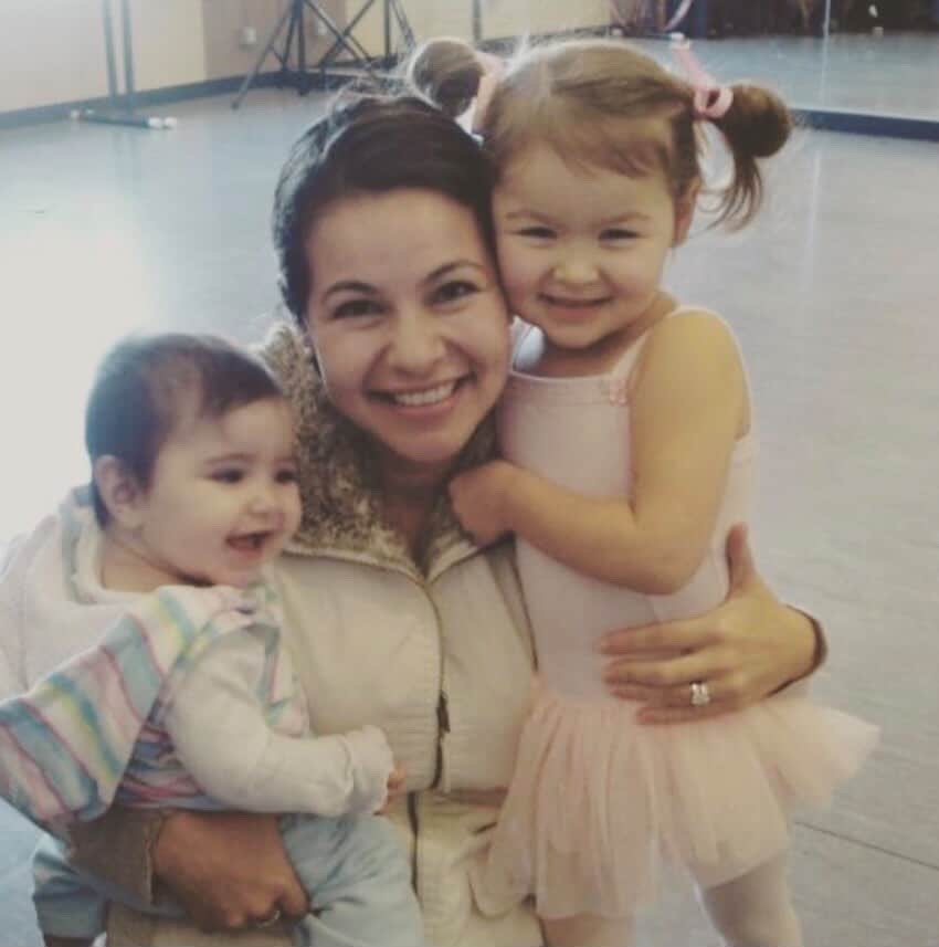 A woman smiling and hugging two young children, one girl with pigtails and the other boy, in an indoor setting.