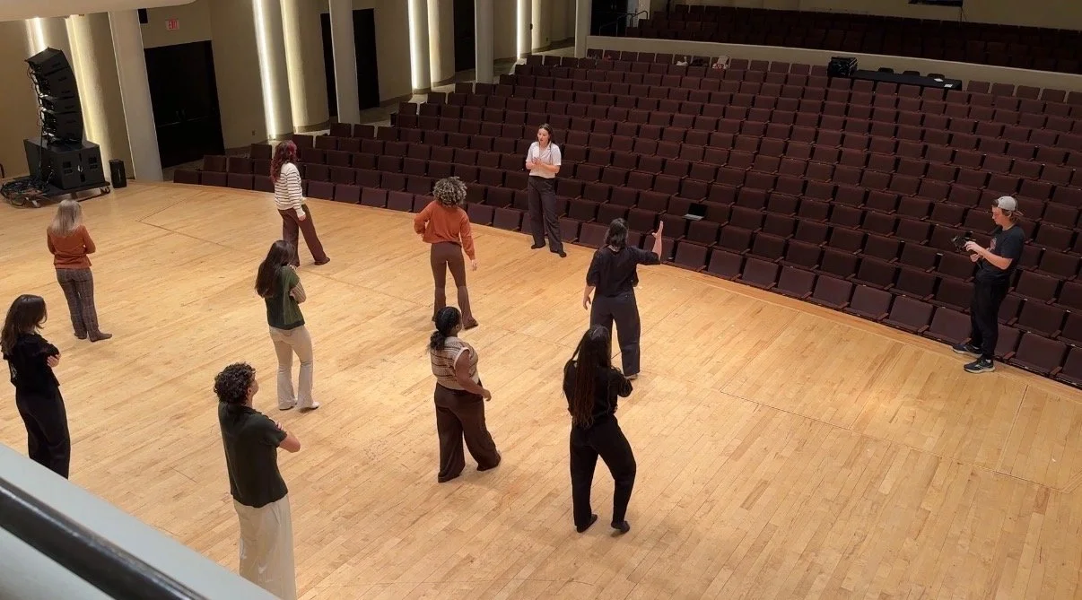 People practicing or rehearsing on a stage in an empty auditorium with rows of theater seats in the background.