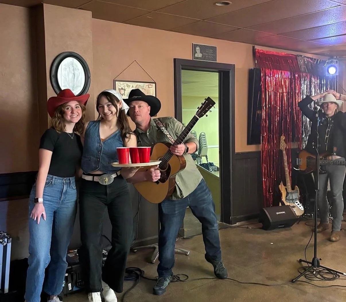 Three people dressed in cowboy and cowgirl attire, with cowboy hats and jeans, stand together at a social gathering. The person in the middle holds a tray with red cups, and the person to their right plays a guitar. In the background, another musician wearing a cowboy hat is near musical equipment and a red curtain.