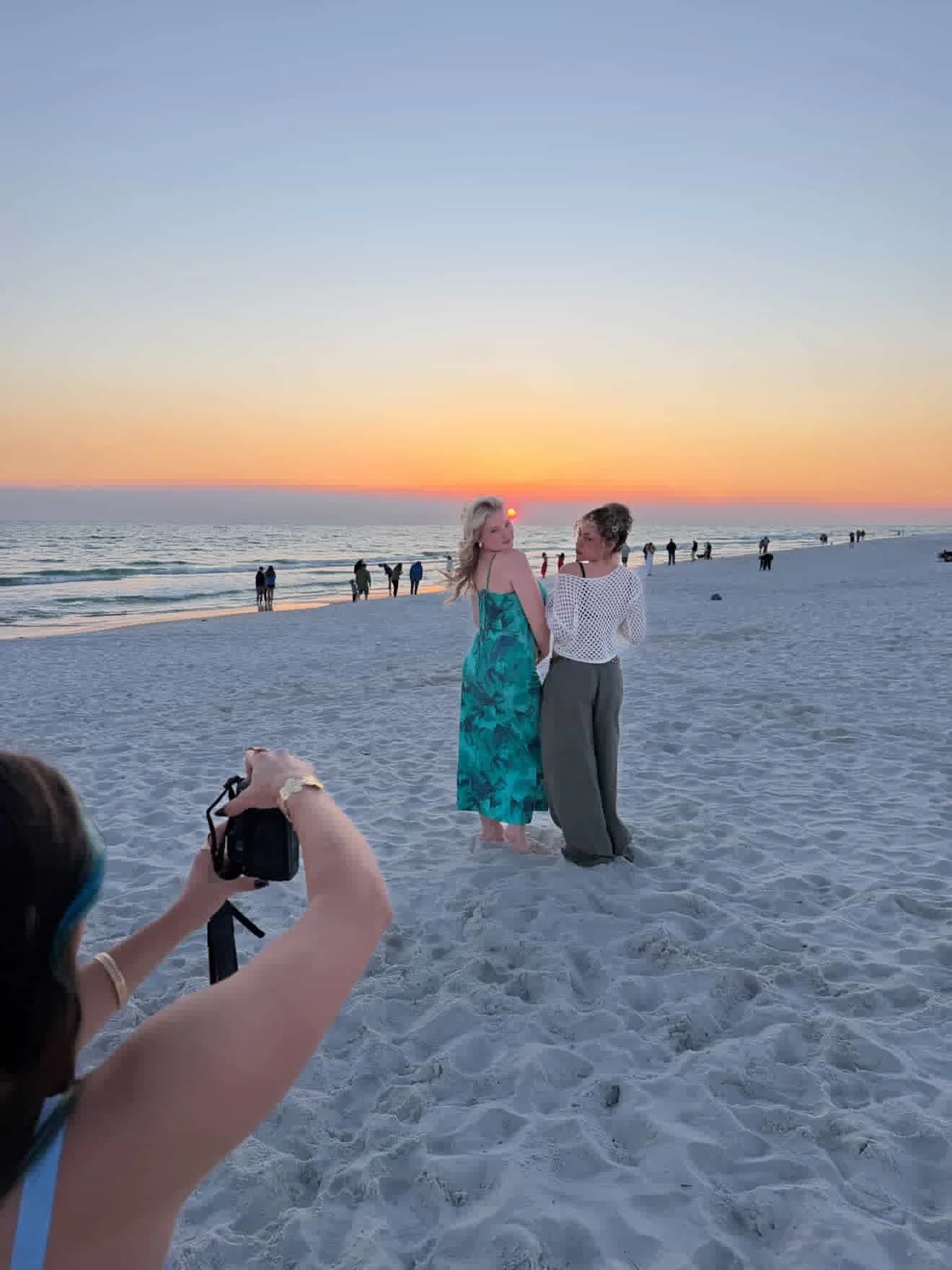 Two women standing on a beach at sunset, posing for a photo while a person in the foreground takes their picture.