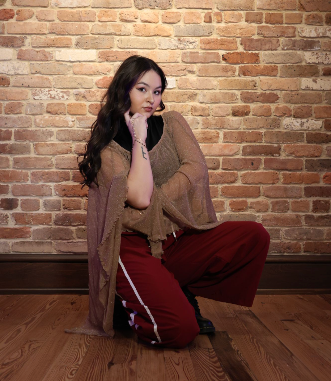 A young woman with long wavy dark hair, kneeling on a wooden floor in front of a brick wall, wearing a sheer gold top, black shirt, and maroon sweatpants with white stripes.