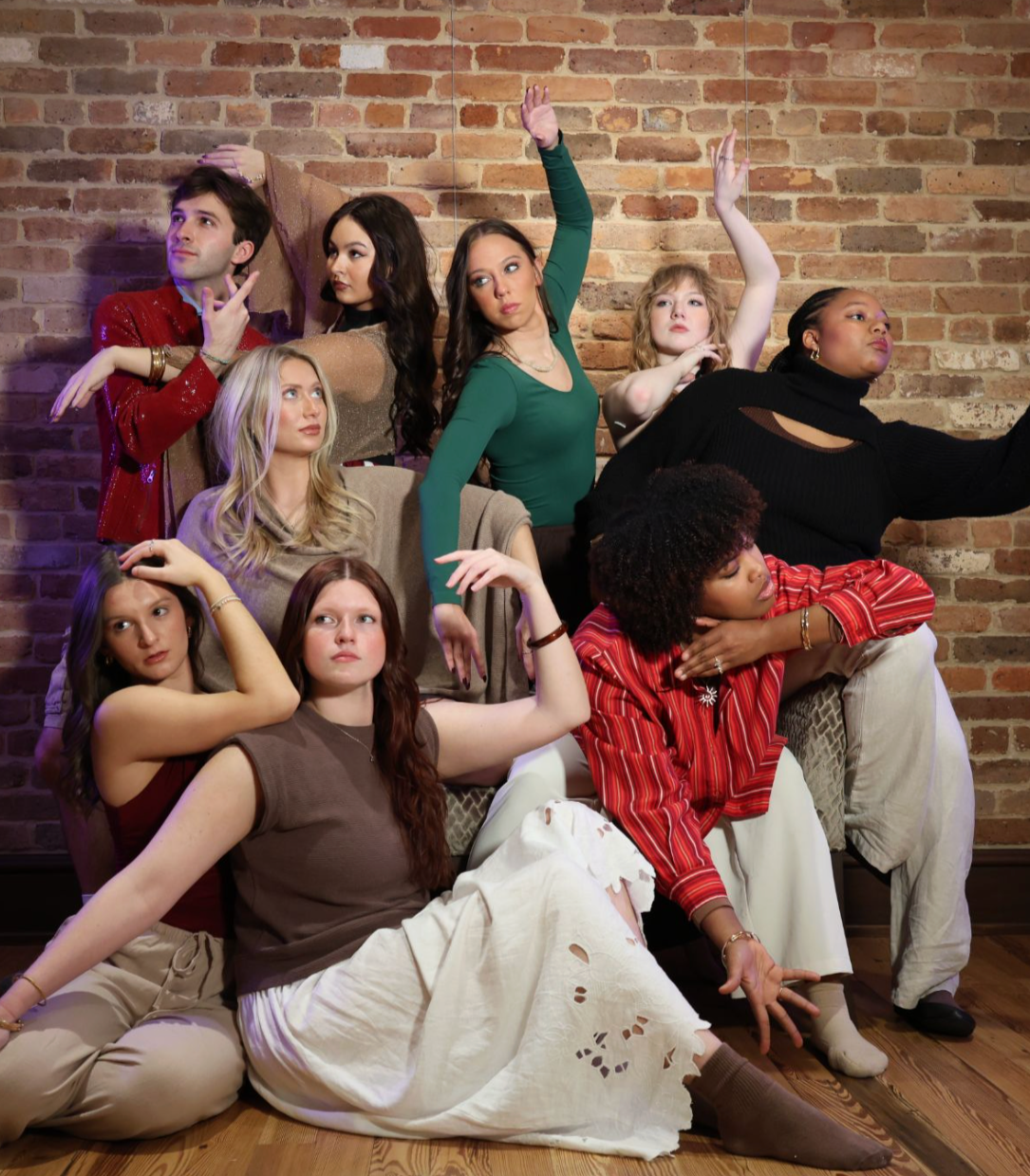 A group of nine diverse young adults posing against a brick wall, some sitting on the floor, others standing or leaning, in a variety of casual and fashionable outfits, striking different poses.