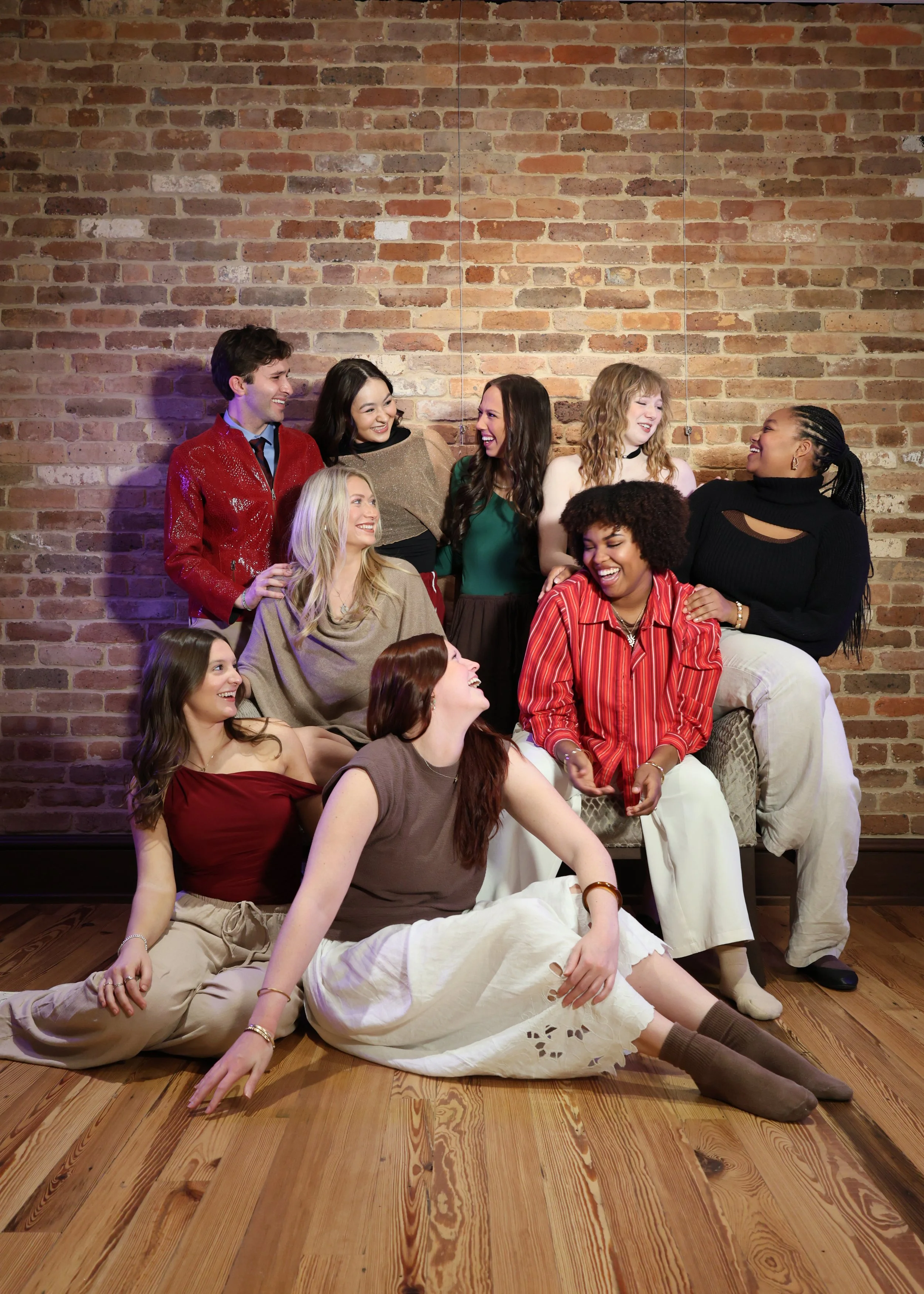 Eight women sitting and standing against a brick wall, laughing and enjoying each other's company.