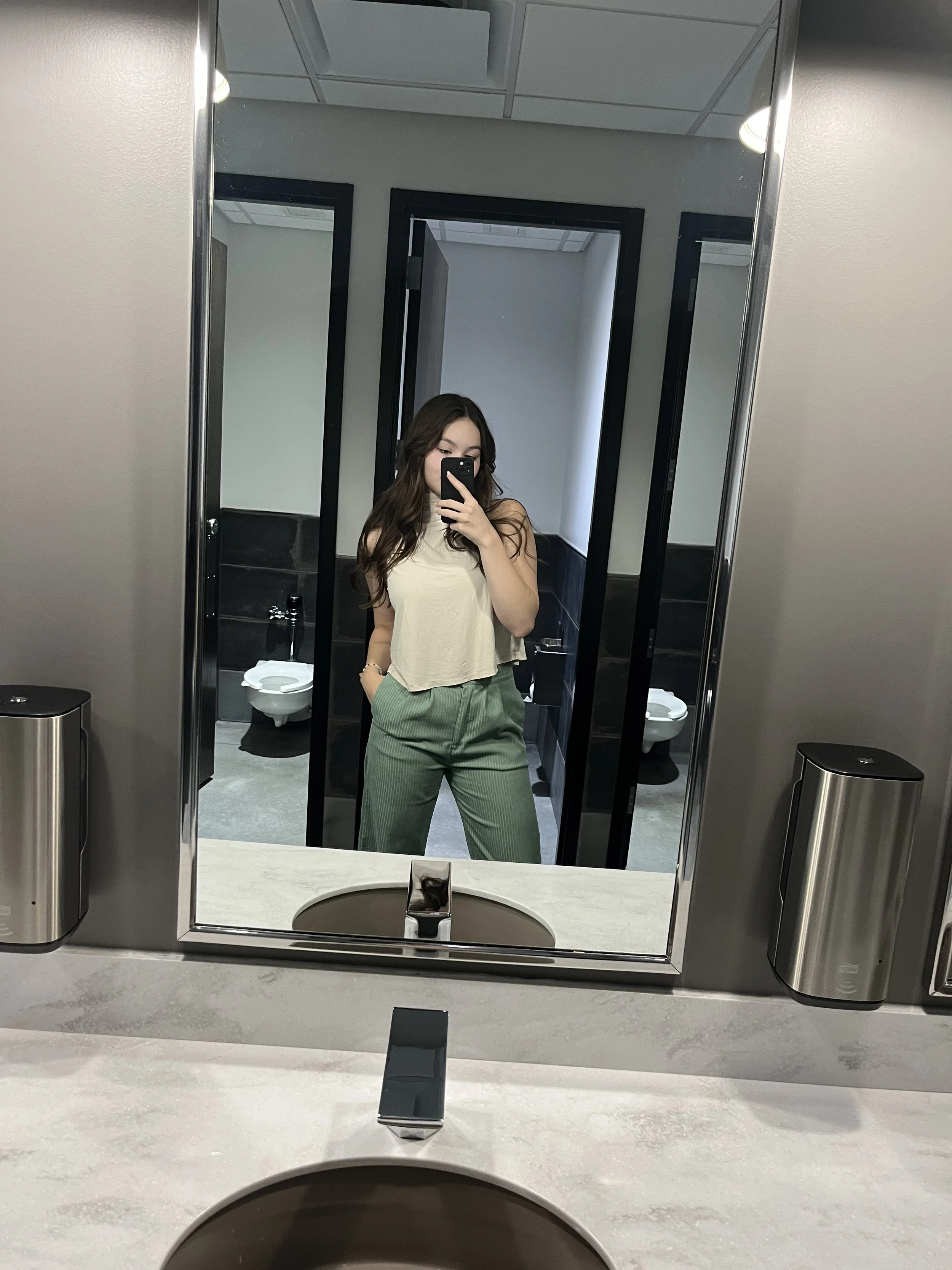 A young woman with long brown hair taking a selfie in a bathroom mirror. She is wearing a cream-colored sleeveless top and green pants. The bathroom has a white sink, a black and white tile wall, and a concealed toilet with a black flush sensor. The mirror is framed in silver, and there are two soap dispensers attached to the wall on either side of the mirror.