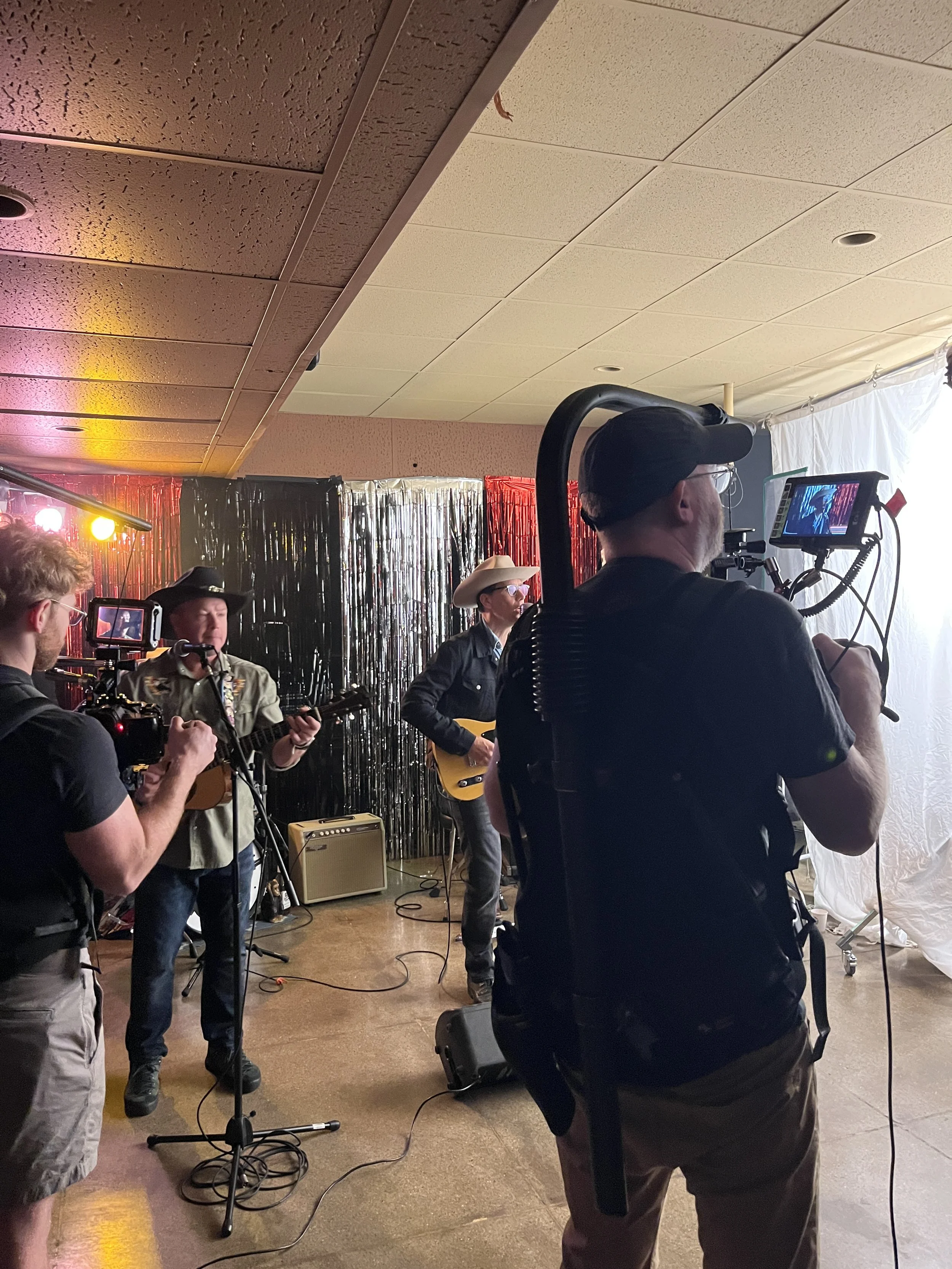 A group of musicians performing on stage with various instruments, including guitars, microphones, and music stands, in a room decorated with metallic streamers and colorful stage lighting.