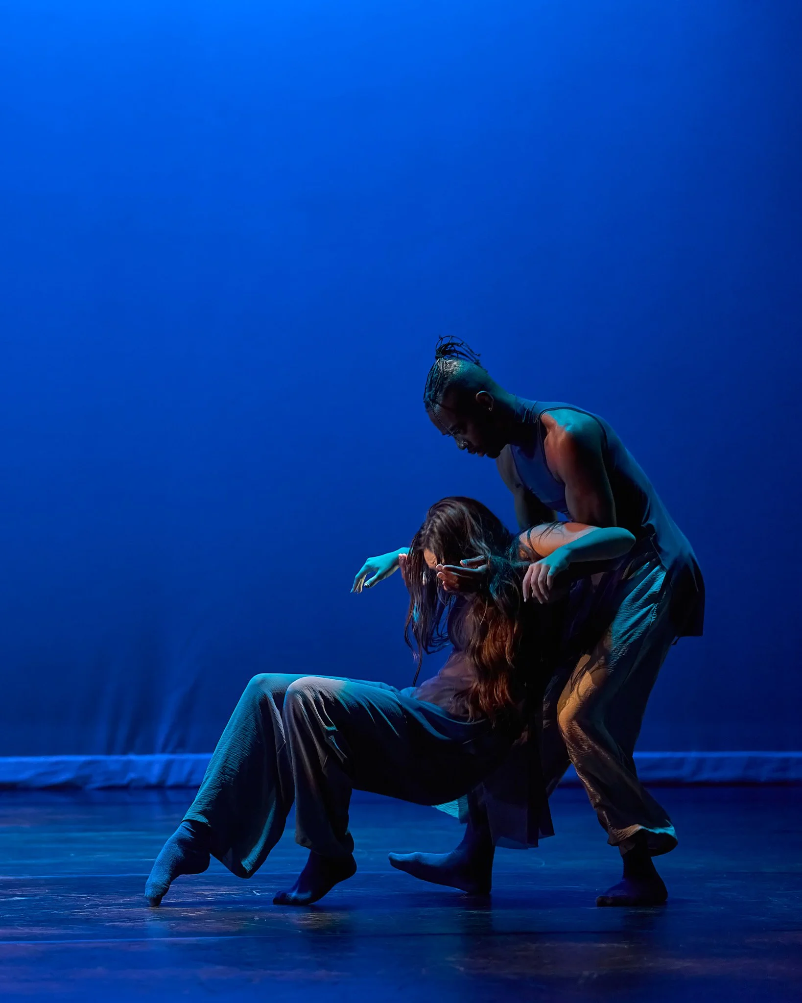 Two performers on stage, one with a mohawk hairstyle and muscular build, and the other with long wavy hair, engaging in a contemporary dance routine with a vivid blue background.