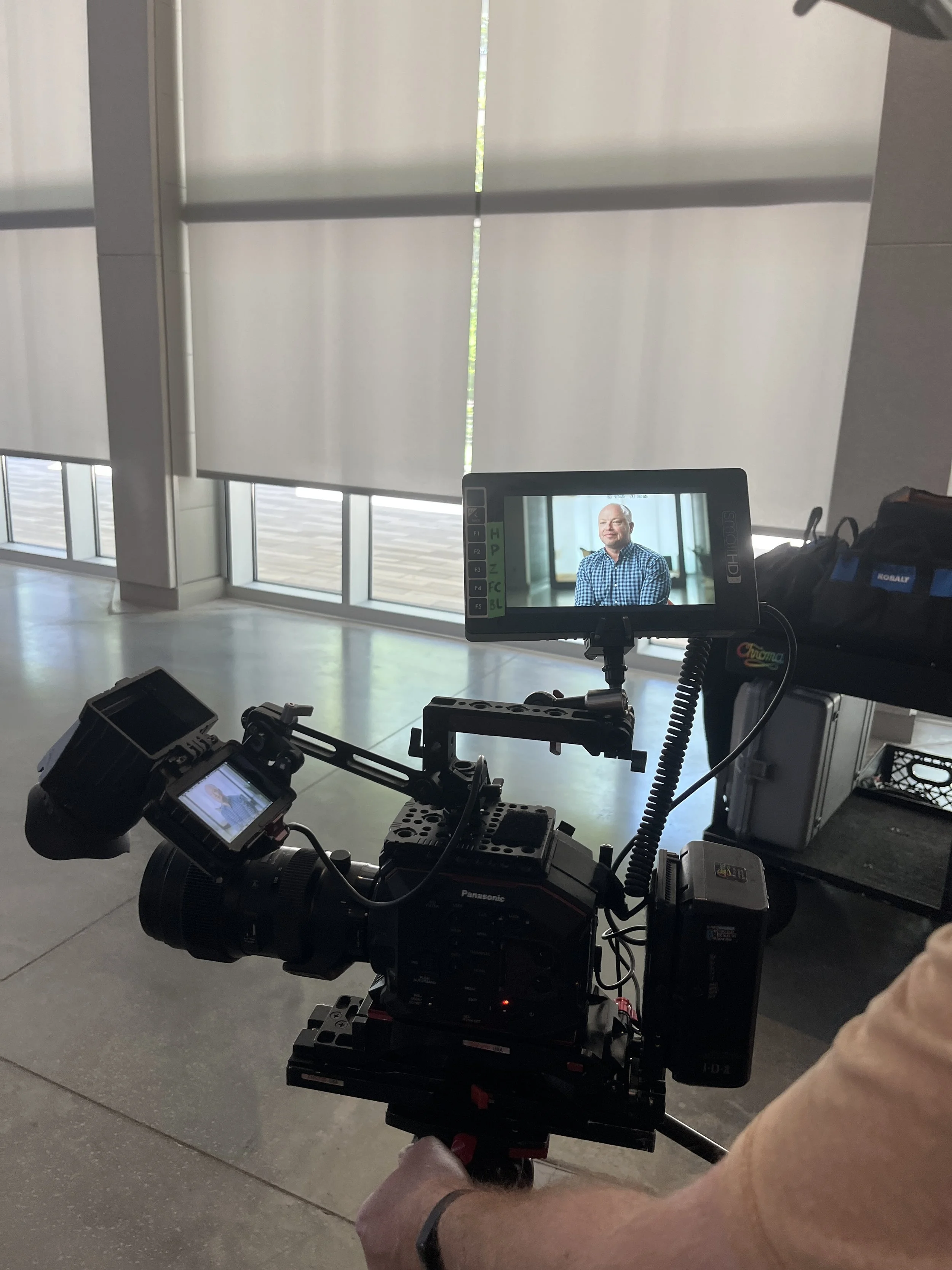 A professional video camera mounted on a stabilizer is capturing an interview or portrait session of a bald man with a beard, wearing a checkered shirt, sitting in an indoor setting with large windows and blinds in the background.