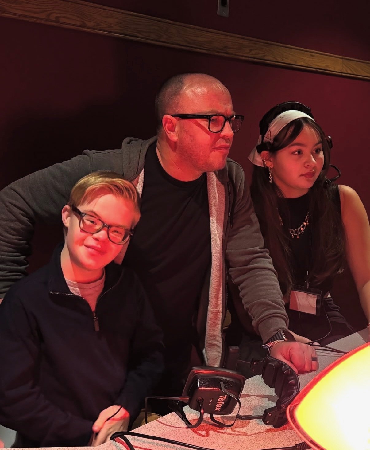 Three people sitting together at a table, with a warm orange glow from a lamp nearby. They are wearing headphones and appear to be in a recording studio or radio station. The person on the left is a young boy with glasses and short hair, smiling. The person in the middle is a bald man with glasses, wearing a hoodie, looking to the side. The person on the right is a woman with long dark hair, wearing earrings, a headscarf, and a black top, also looking to the side.