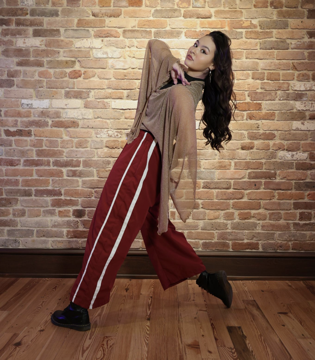 A woman poses against a brick wall, wearing red pants with white stripes, a sparkly gold top, and black boots.