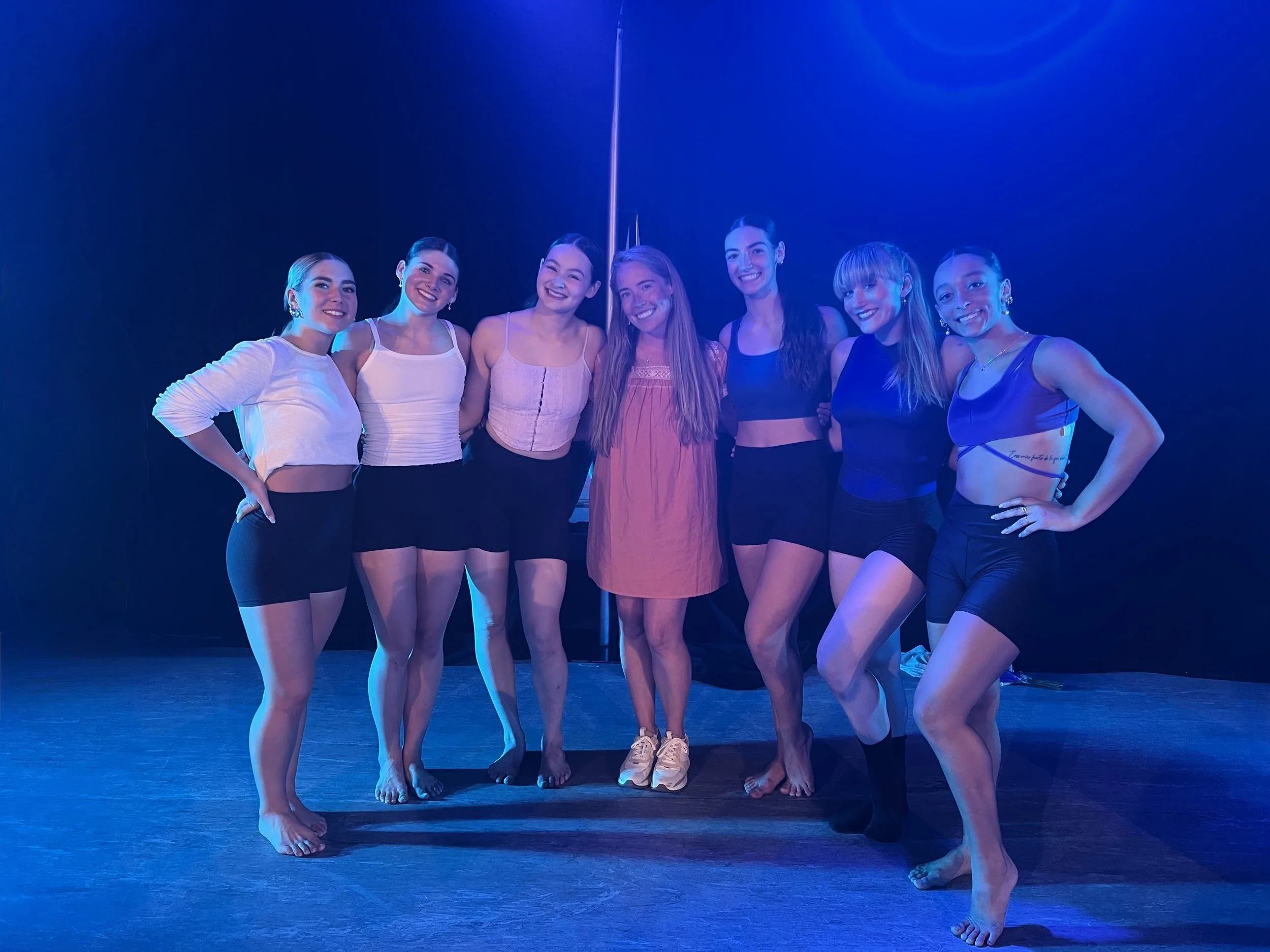 Group of seven young women posing together on a dance stage under blue lighting, smiling at the camera.