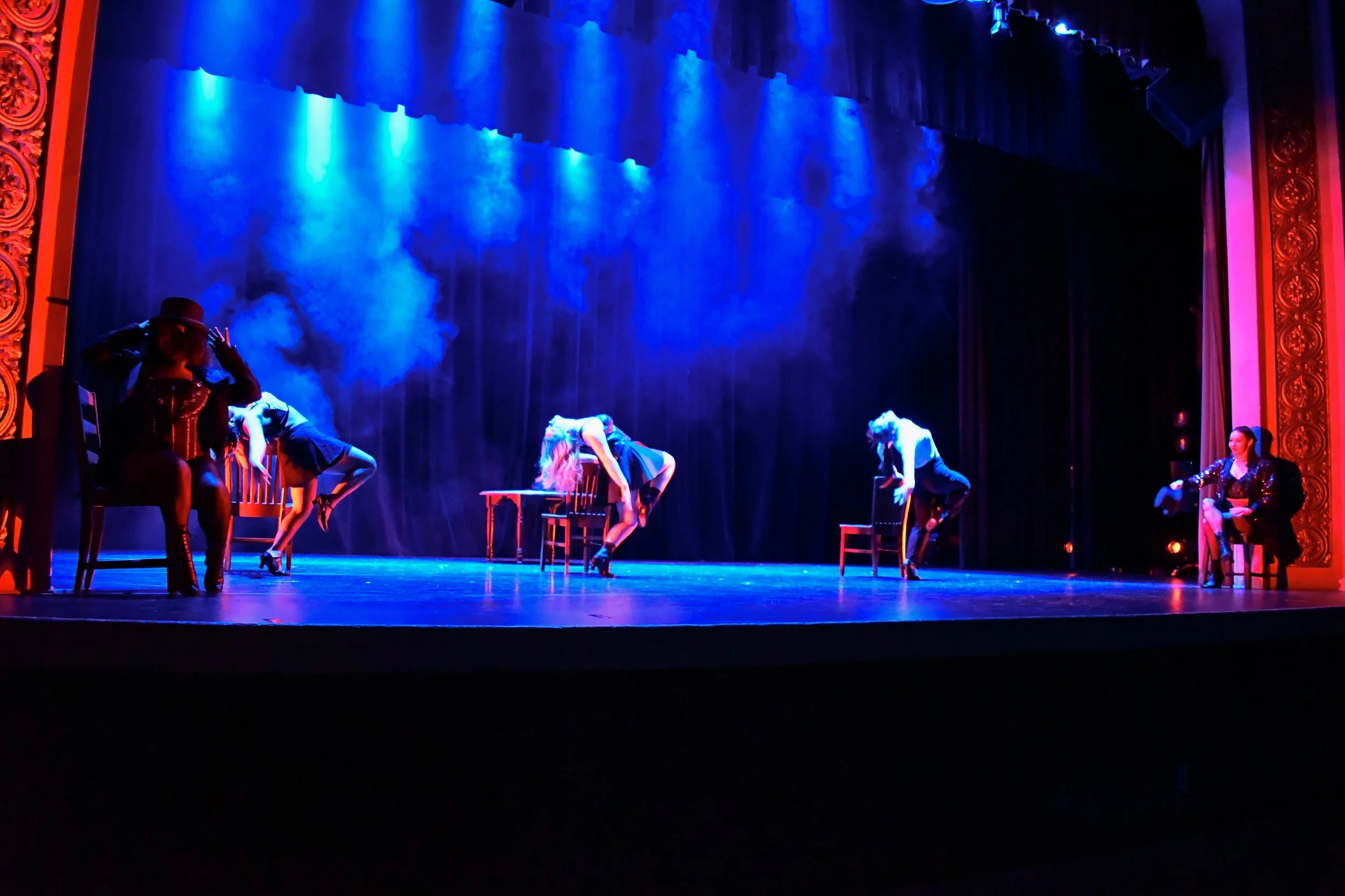 Stage with five performers, some bending forward on chairs, illuminated by blue and purple lighting, ornate gold and red theater framing.
