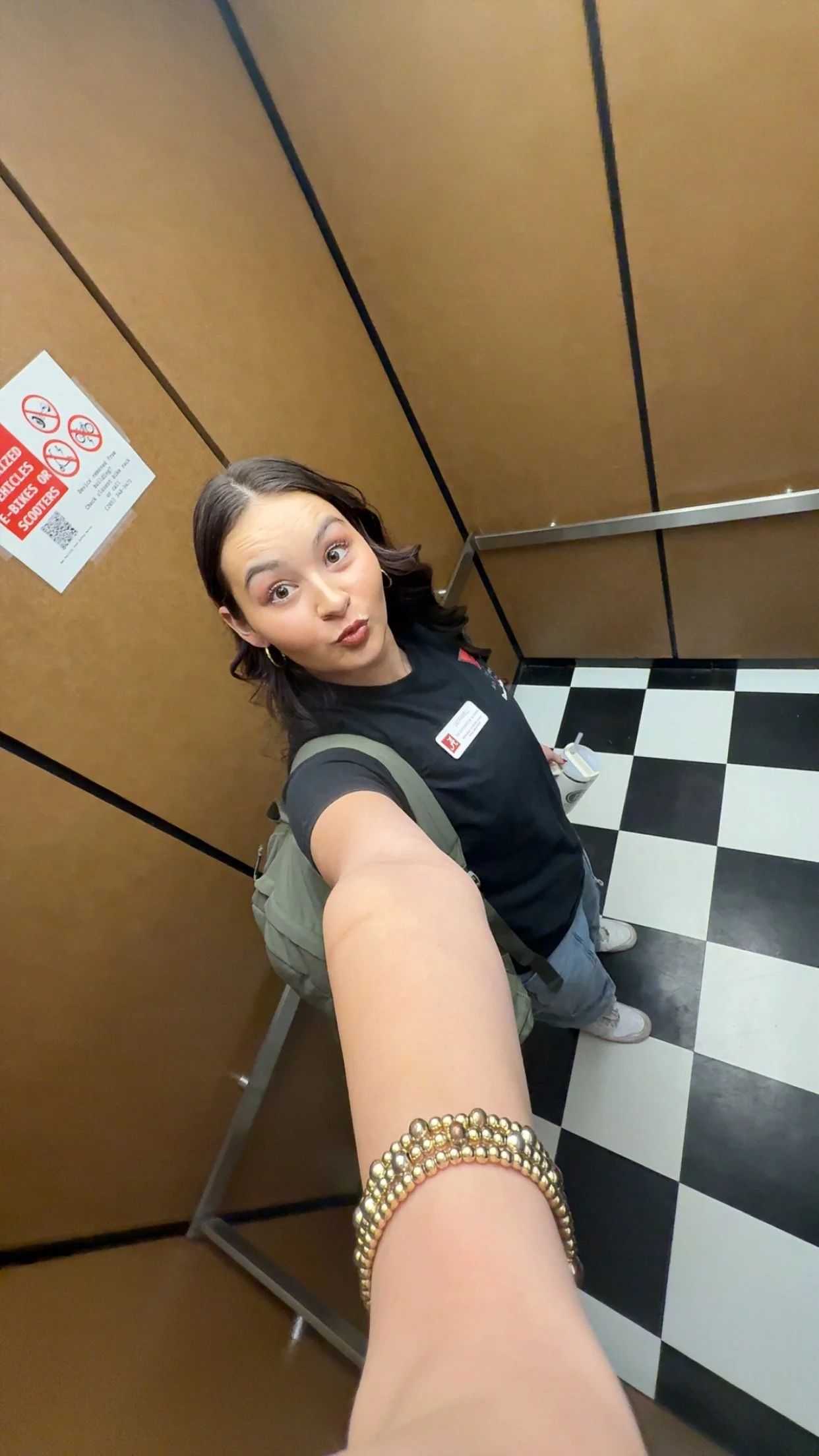 A woman taking a selfie in an elevator, making a playful face, with a brown pantry and black-and-white checkered floor visible.
