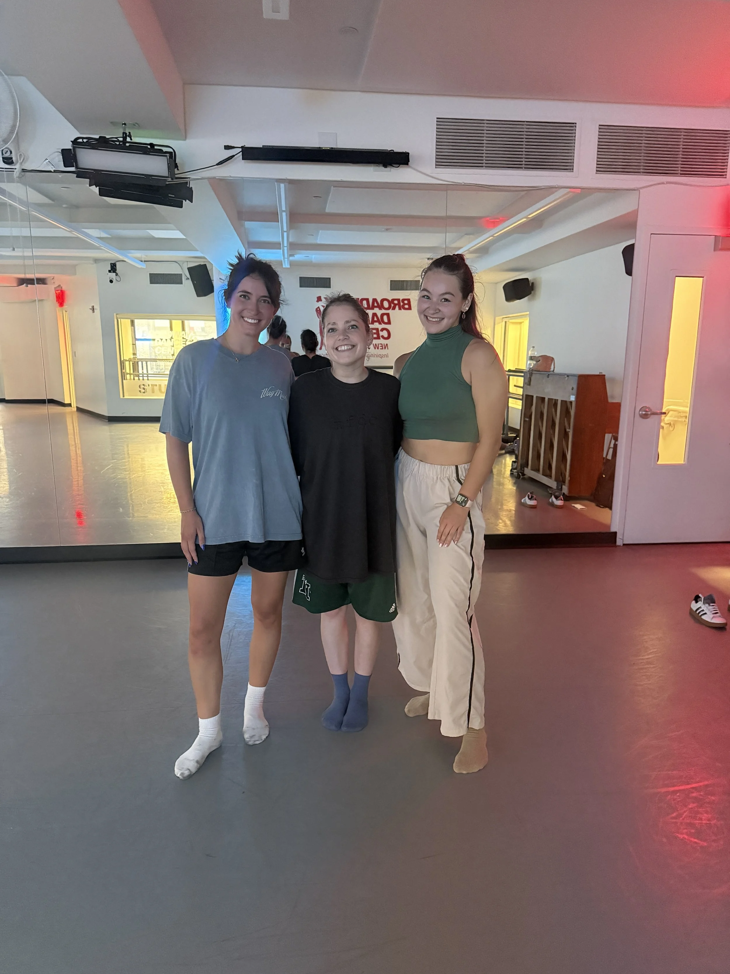 Three young women standing together indoors, smiling for the camera. One is wearing a blue t-shirt and black shorts, another is in a black T-shirt and green shorts, and the third woman is wearing a green sleeveless crop top with beige pants.
