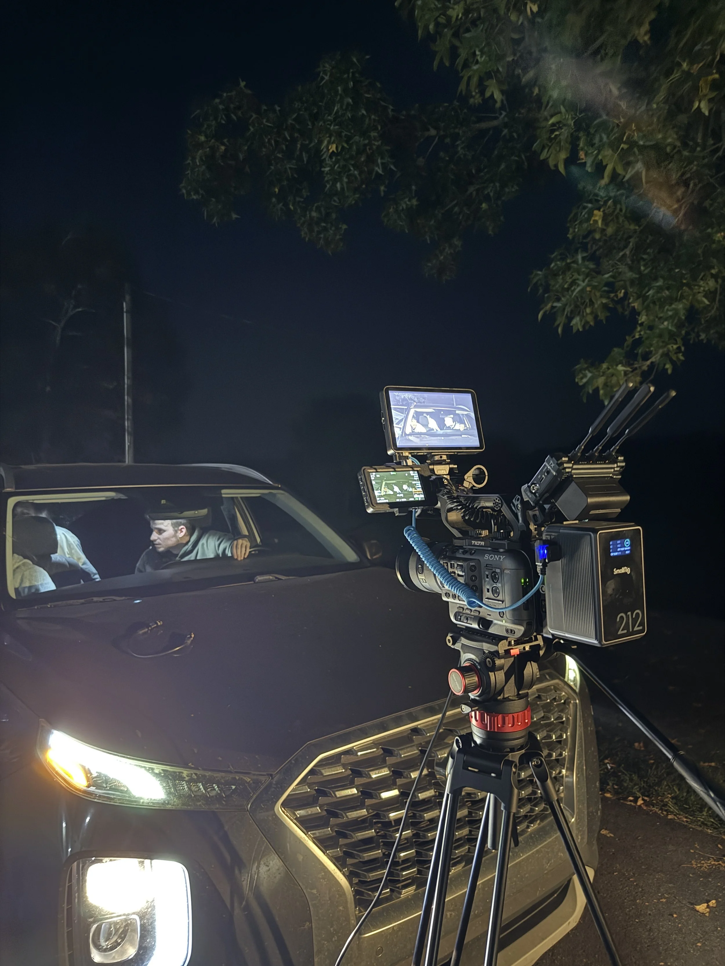 Night scene showing a camera on a tripod facing a black car with illuminated headlights. A crew member is inside the car, and filming equipment, including monitors, is set up around the vehicle. There are trees and power lines in the background.