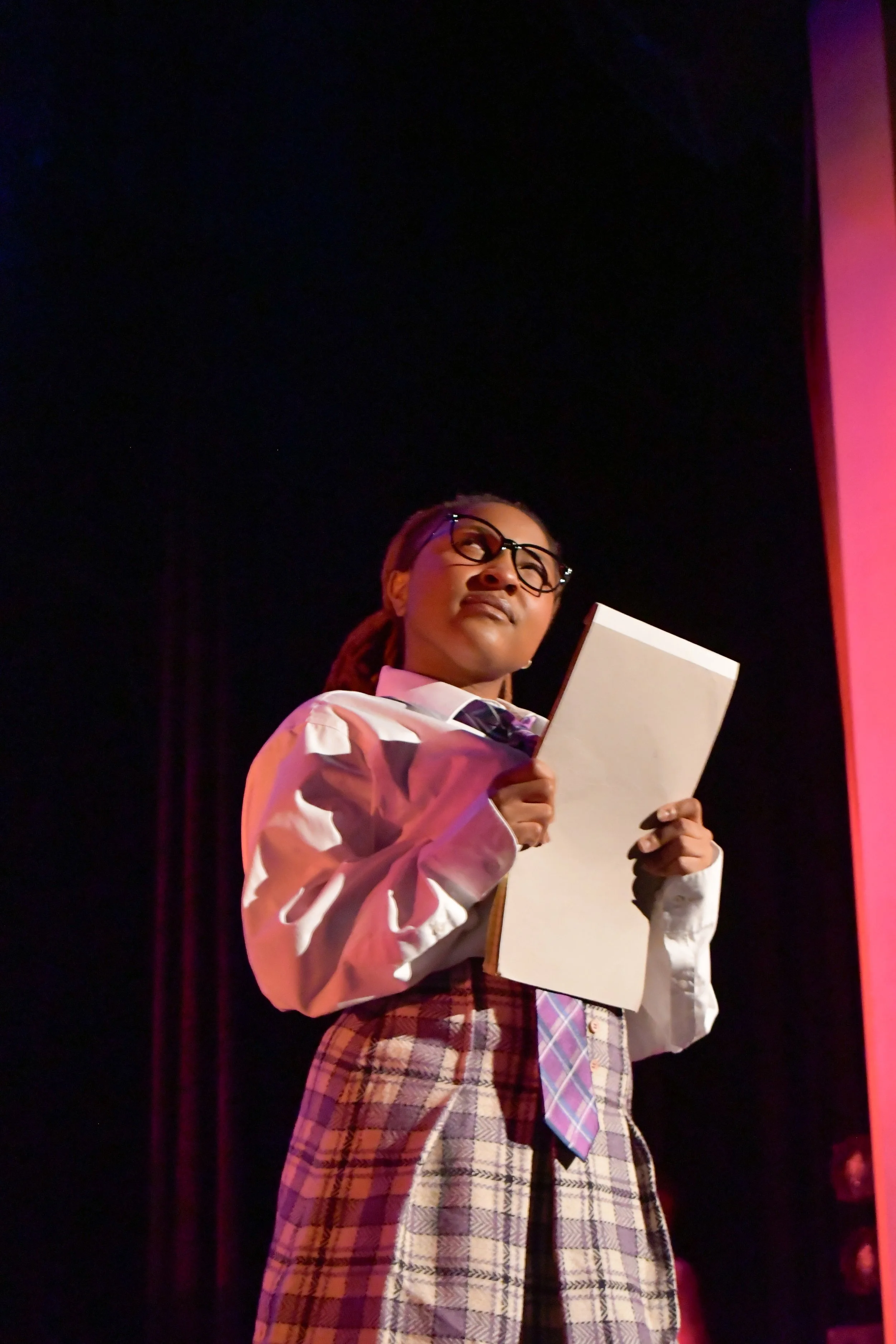 A young woman with glasses and a plaid skirt, holding a notebook, standing against a dark background with pink lighting.