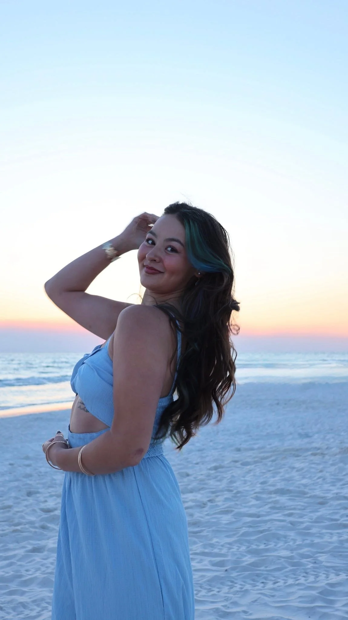 A woman standing on the beach during sunset, smiling at the camera, with her hair partially dyed blue, wearing a light blue dress.