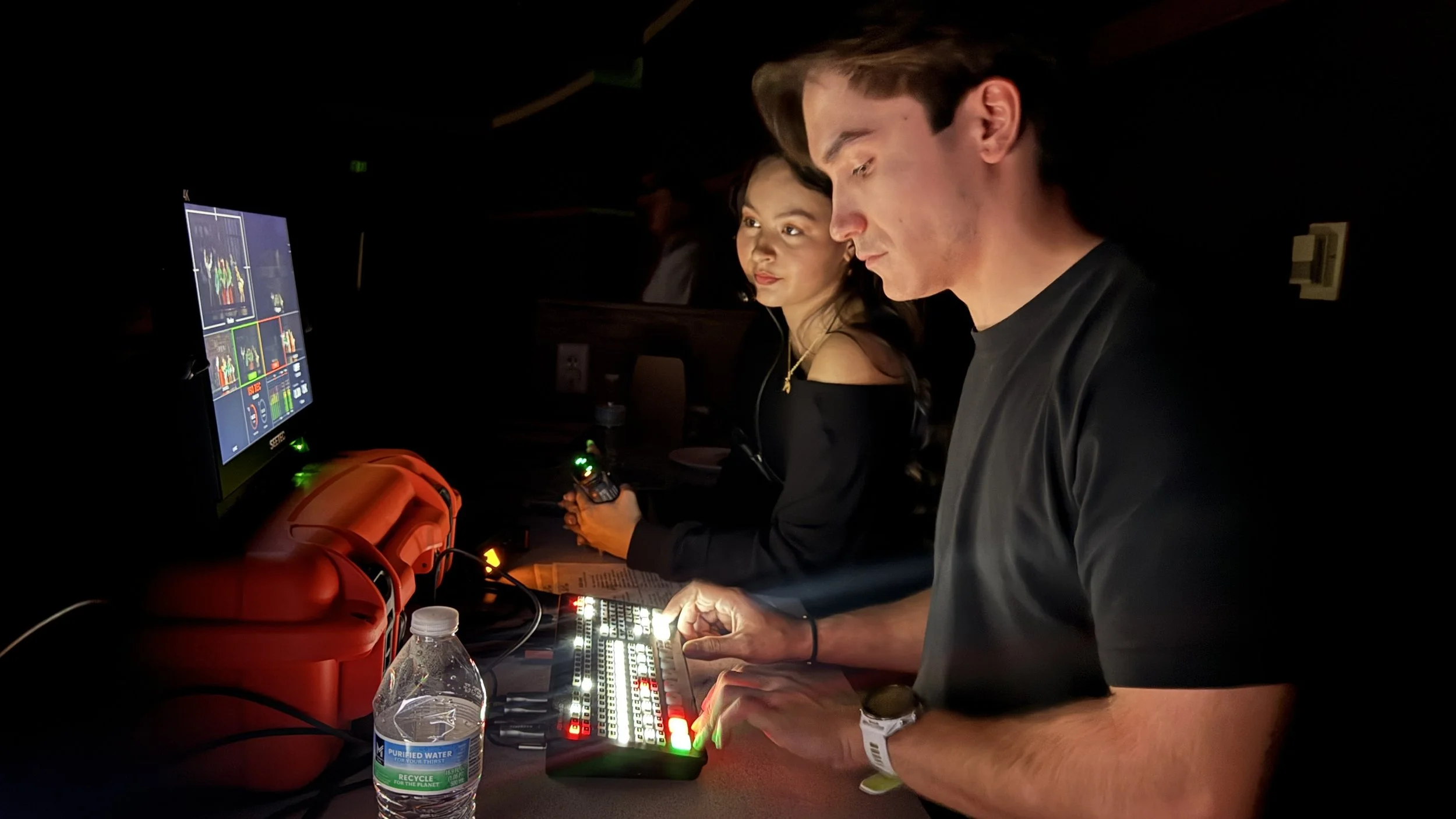 People operating sound and lighting equipment at an event in a dark environment, with a computer monitor displaying sound mixing controls.