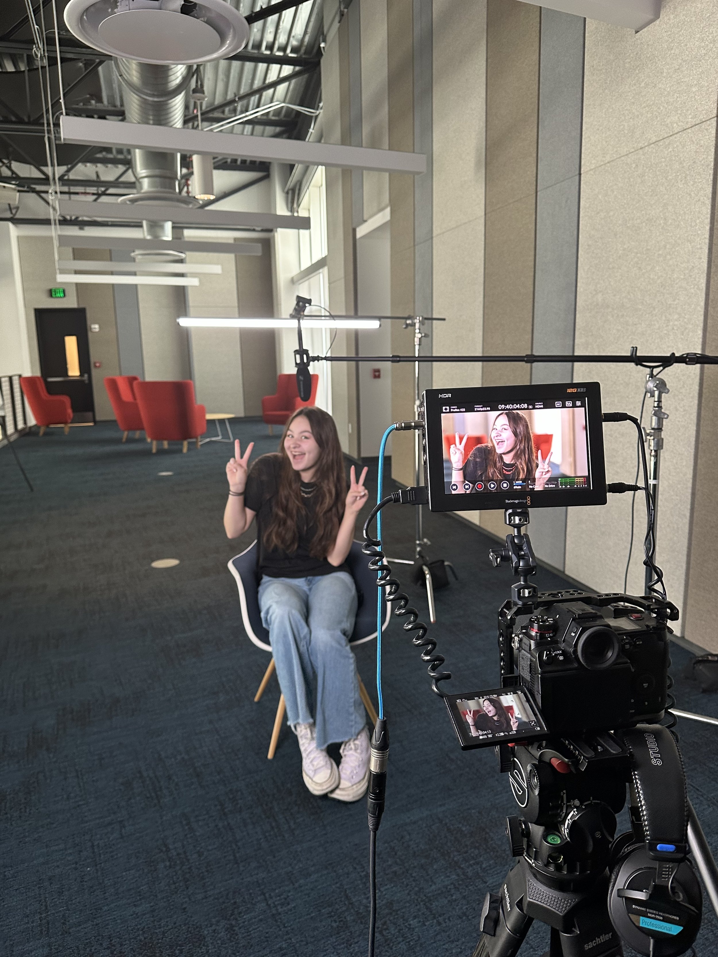 A young woman sitting in a modern studio with red chairs in the background, making peace signs and smiling for a video recording on a professional camera. The studio has industrial ceiling elements and large windows letting in natural light.