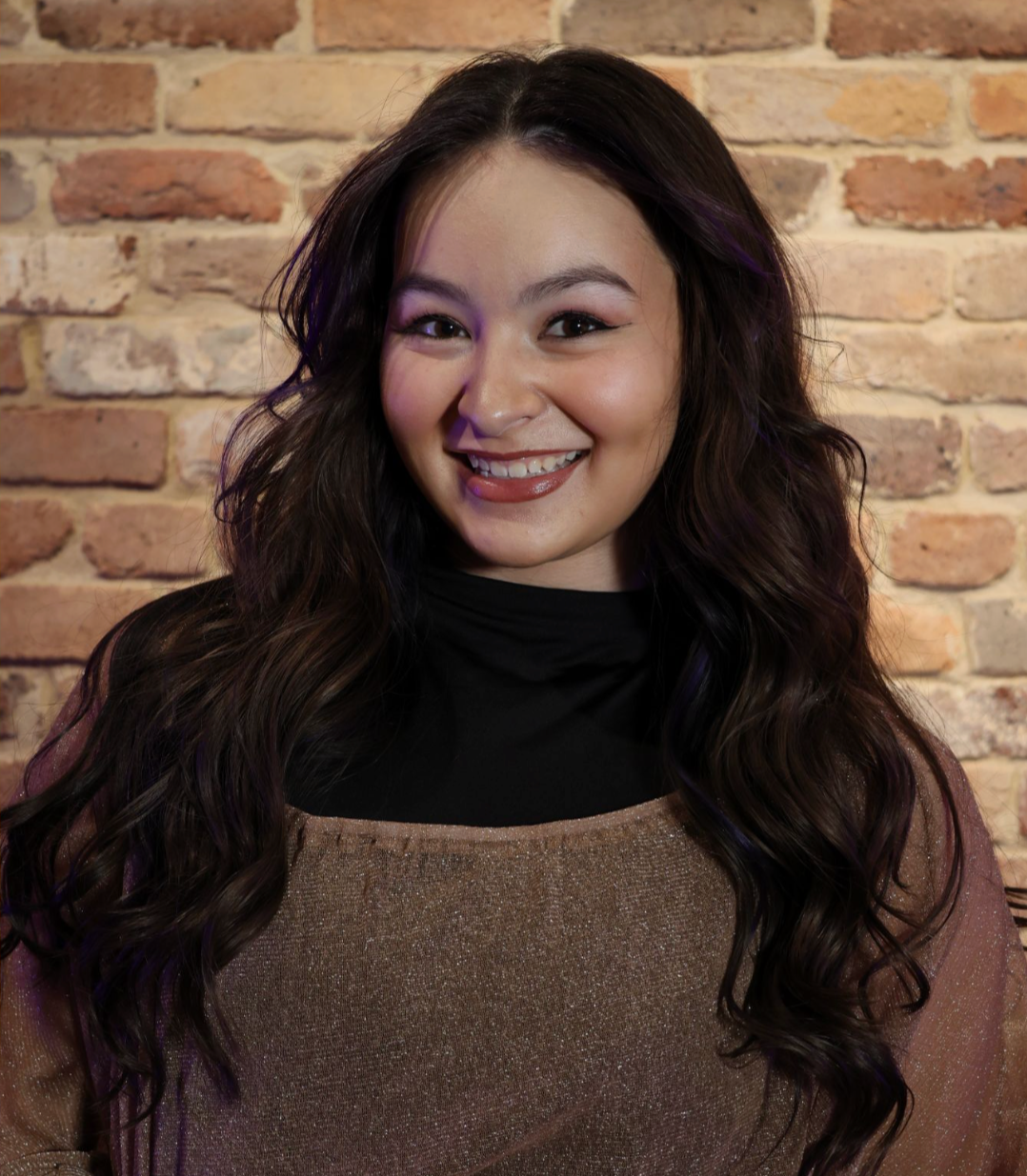 A smiling woman with long wavy dark hair, wearing a black turtleneck and a shimmery top, standing in front of a brick wall.