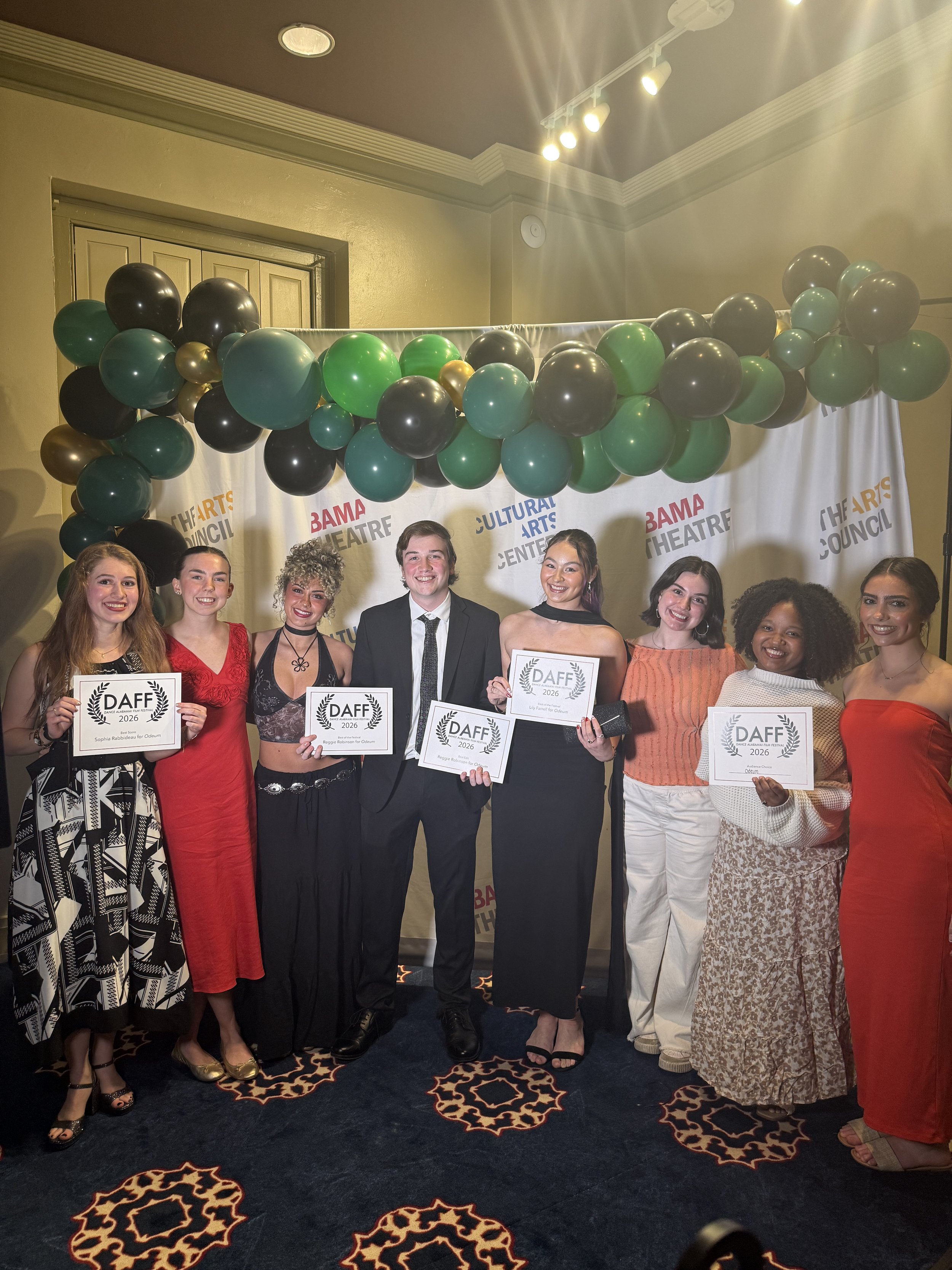 Group of people at an award ceremony holding certificates with a backdrop reading 'BAMA THEATRE CULTURAL ARTS CENTER'. The individuals are dressed in formal and semi-formal attire and are smiling. There is a balloon decoration overhead.