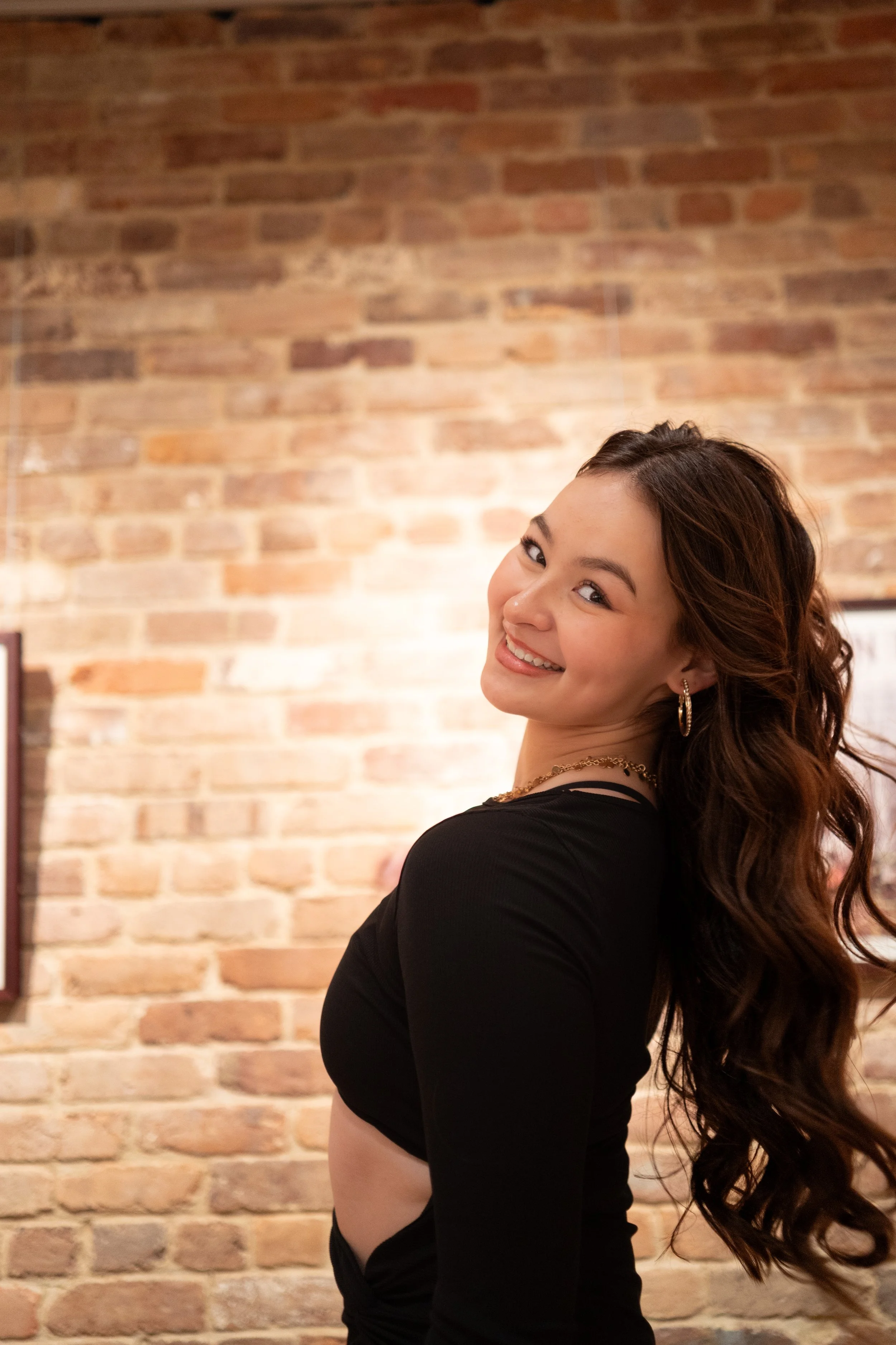 A woman with long, wavy brown hair smiling at the camera, wearing a black long-sleeve top, earrings, and a necklace, standing in front of a brick wall.