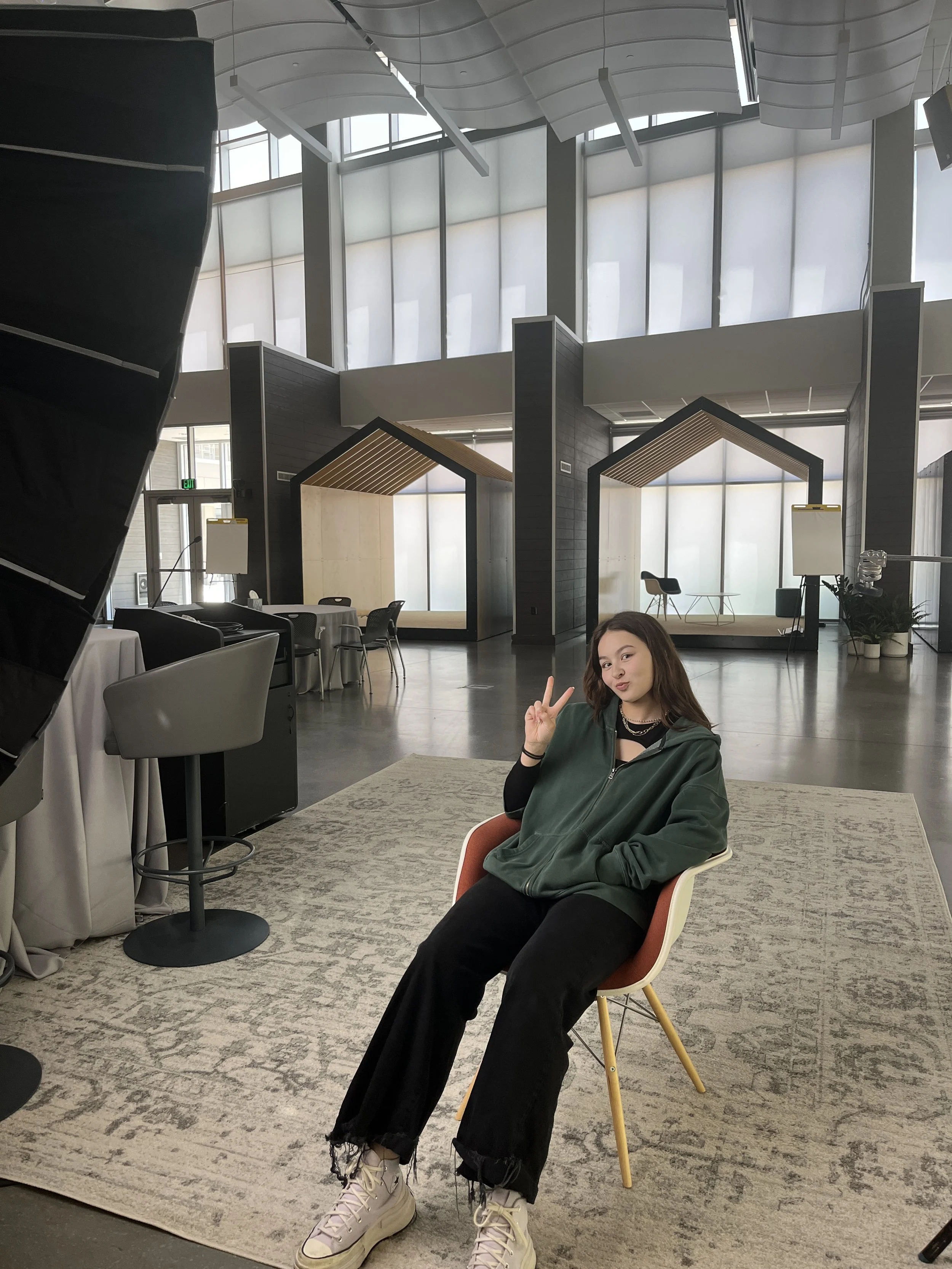 Young woman with brown hair sitting on a pink and yellow chair in an open space with modern architectural design, large windows, and a stage in the background. She is making a peace sign with her right hand and wearing a dark green jacket, black pants with frayed hems, and beige sneakers.
