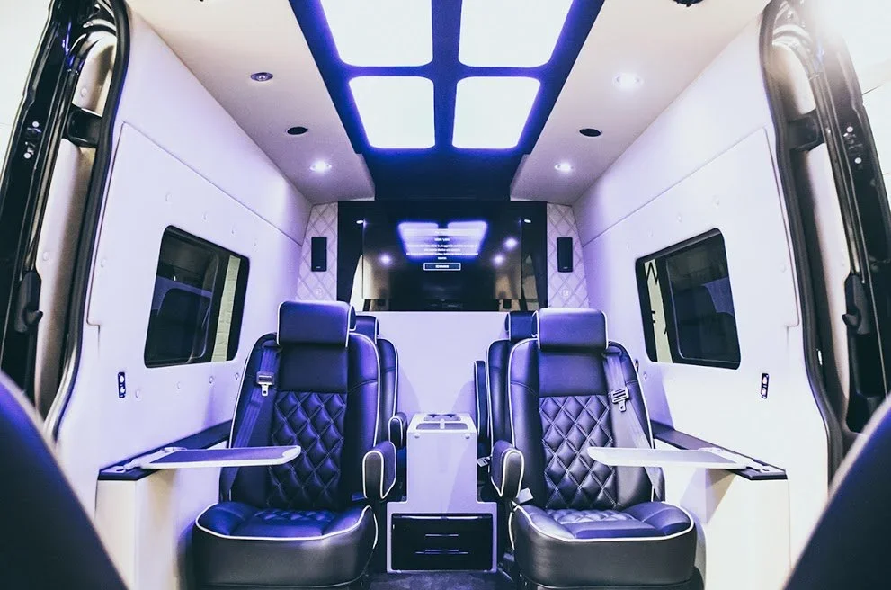 Interior of a luxury van or mobile office with two black leather and quilted seats facing forward, small fold-out tables, large windows, and modern ceiling lighting.