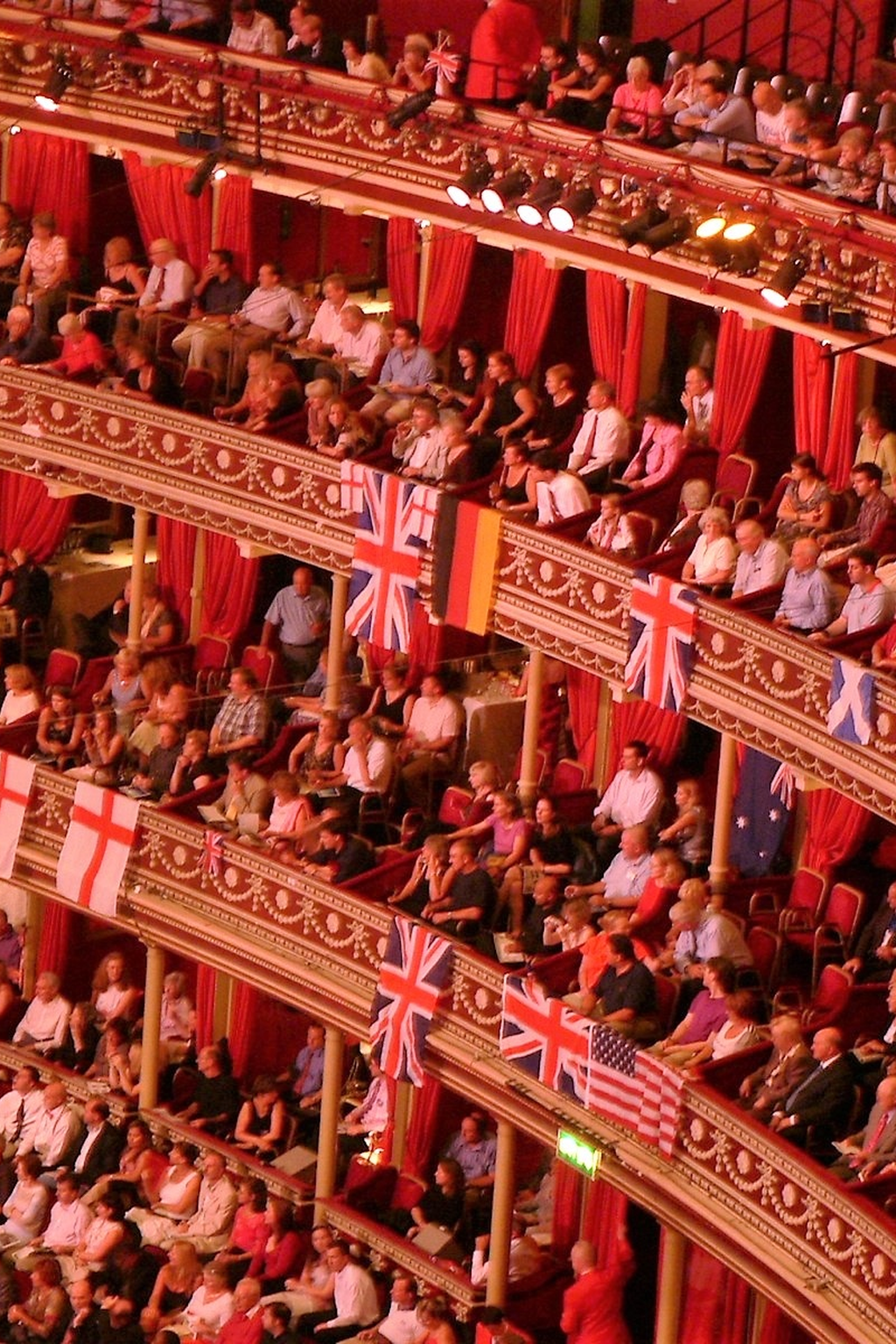 Audience members seated in a theater with ornate red curtains, and British, German, and American flags draped along the tiers.