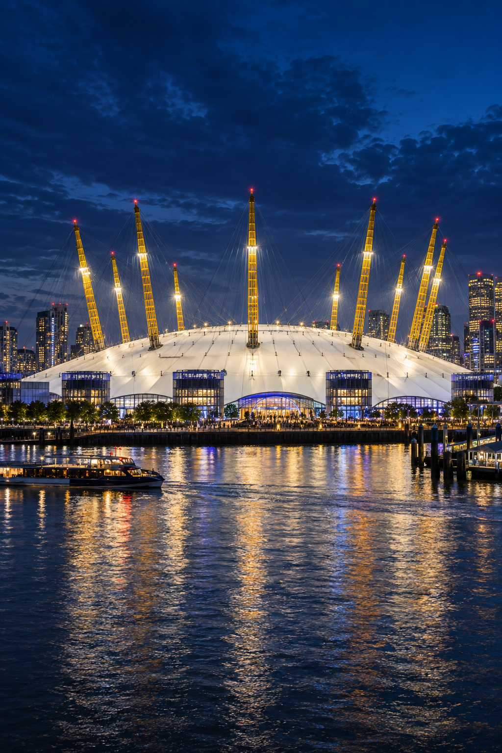 Nighttime cityscape featuring the O2 Arena in Lodon with its illuminated yellow supports and a boat sailing on a reflective water body in front.