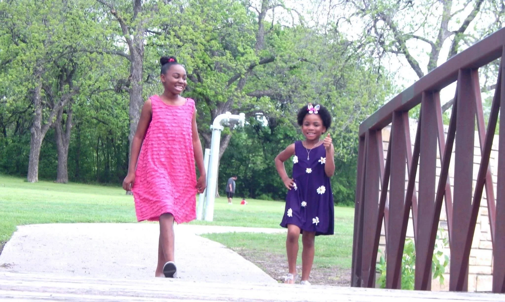 Two girls walking outdoors on a park sidewalk. One girl in a pink dress, the other in a dark blue dress with white flower patterns, smiling and giving a thumbs-up. There are trees and a wooden railing in the background.
