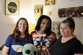 Three women smiling indoors, with decorative wall art, and a plush toy resembling Baby Yoda in front.